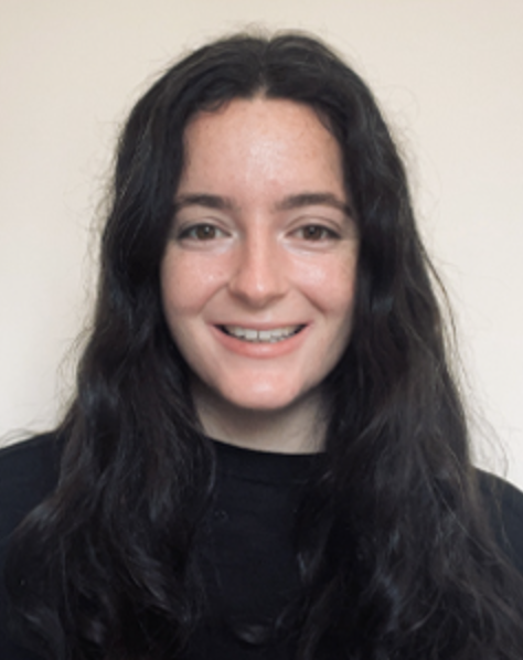 A young woman with long, wavy dark hair smiling in front of a plain light-colored wall.