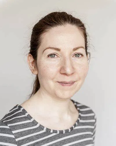 A woman with red hair pulled back, wearing a gray and white striped shirt, smiling lightly at the camera against a plain light background.