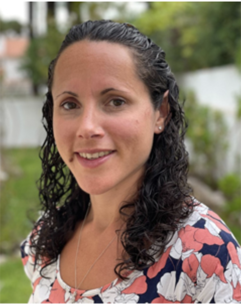 A woman with dark curly hair smiling outdoors, wearing a floral blouse with a garden background.