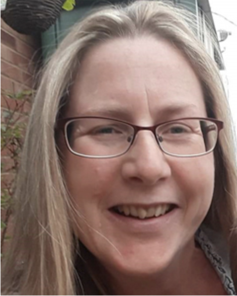 Close-up of a woman with long blonde hair, glasses, smiling outdoors with a brick wall and some greenery behind her.