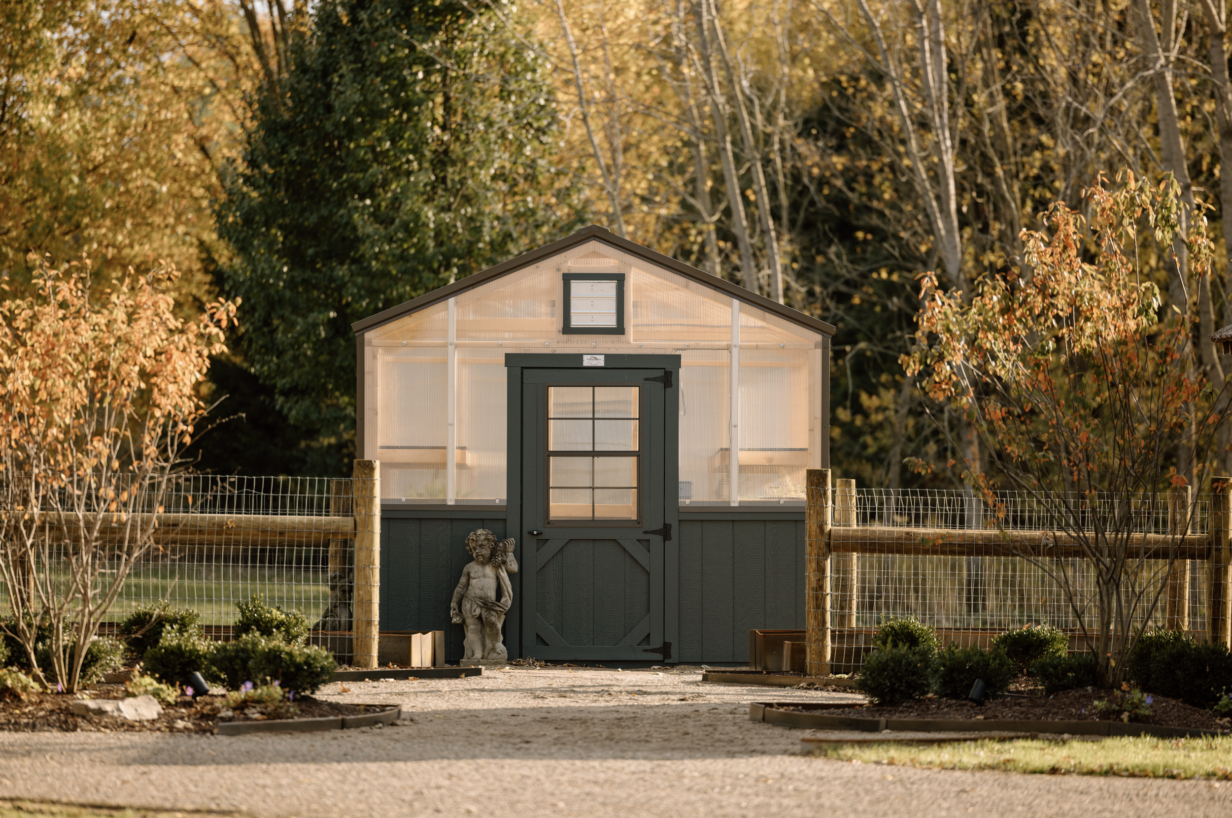A small greenhouse with a dark green door and trim, surrounded by a garden with plants and small trees, trees in fall foliage in the background.