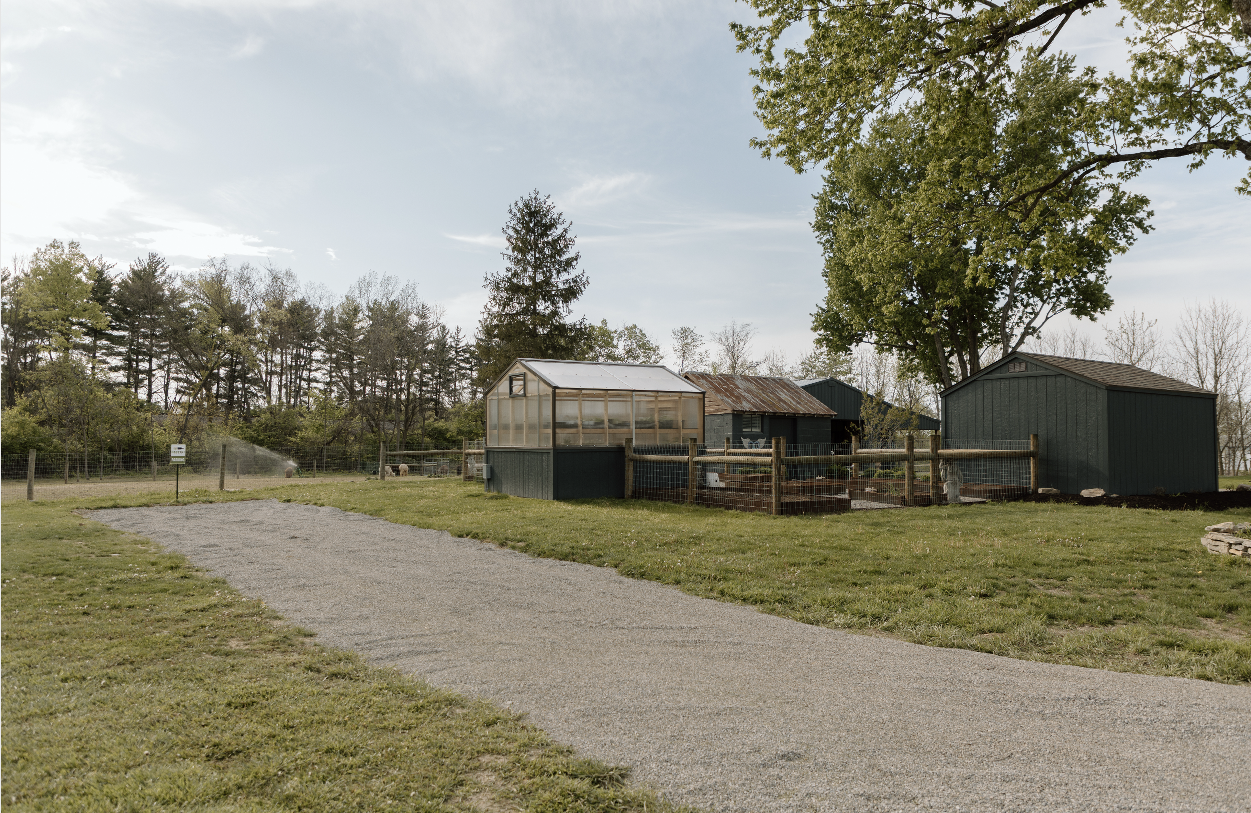 A rural scene featuring a greenhouse, farm buildings, and sheep in a fenced area, surrounded by trees under a partly cloudy sky in Delaware, Ohio