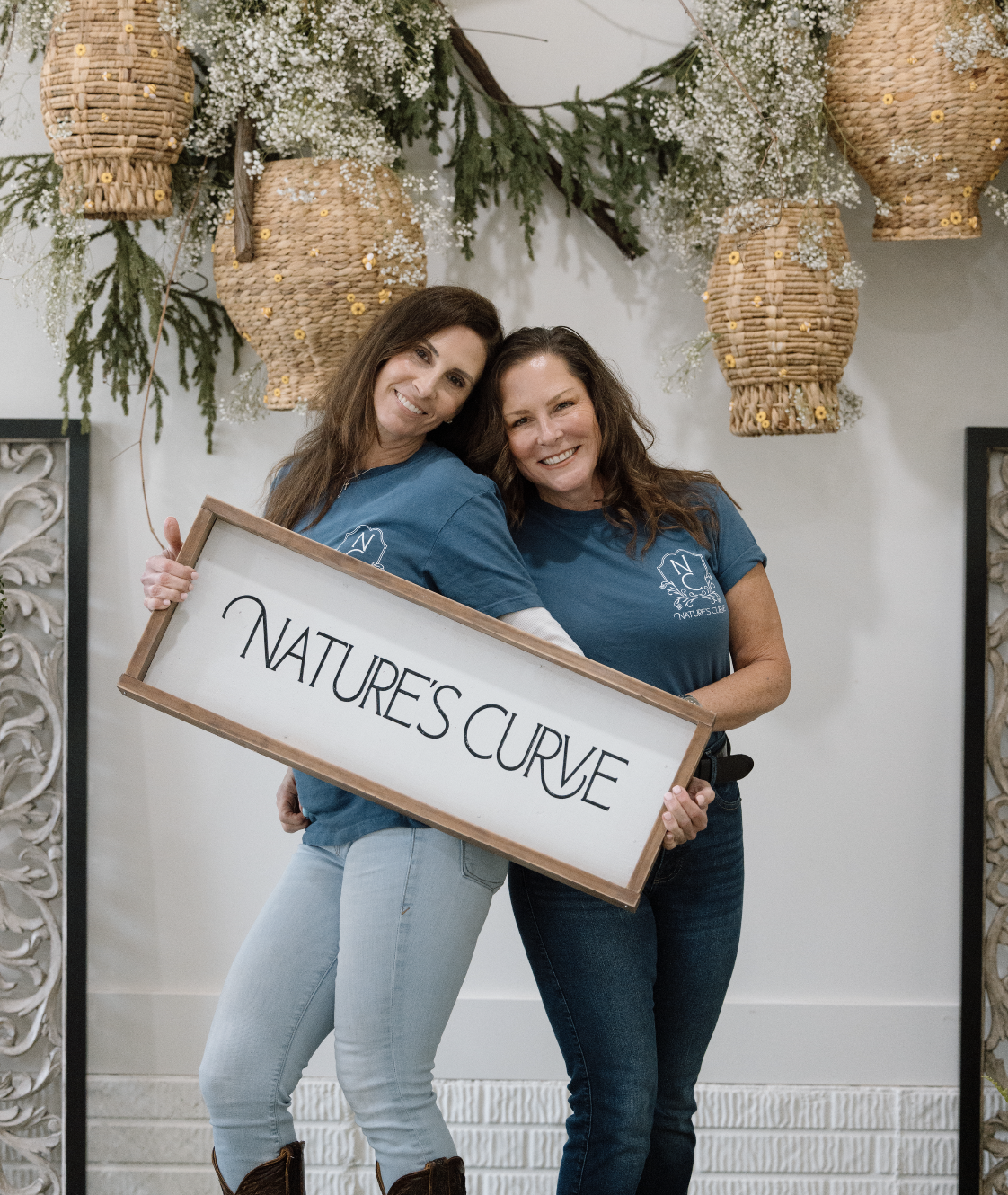Two women smiling and holding a sign that says "Nature's Curve" in front of hanging wicker lanterns and floral decorations in Delaware, Ohio.