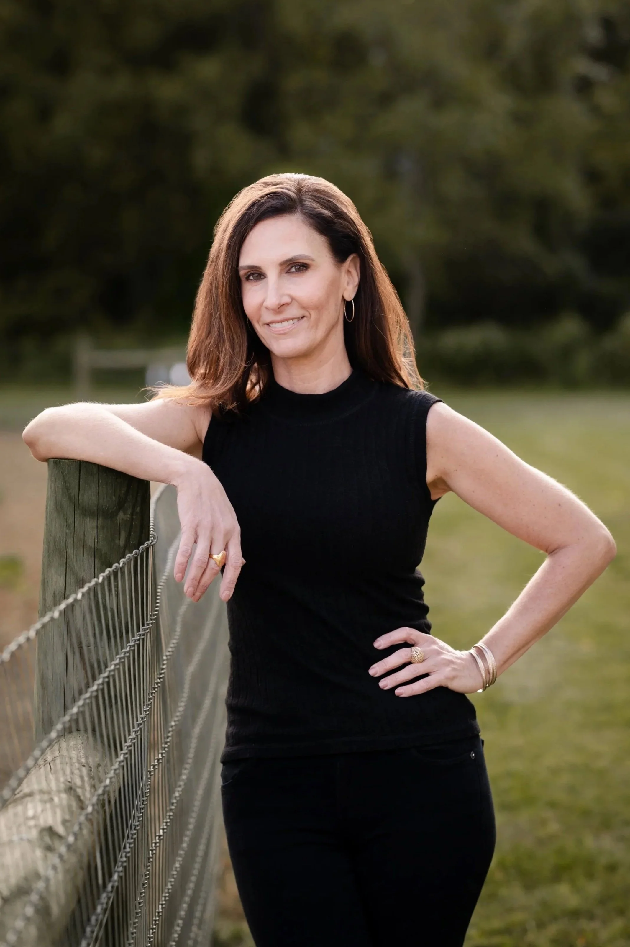 Jaimie Calicut,  the owner of Nature's Curve, seen with brown hair in a black sleeveless top and black pants, leaning on a wooden fence outdoors, smiling at the camera.
