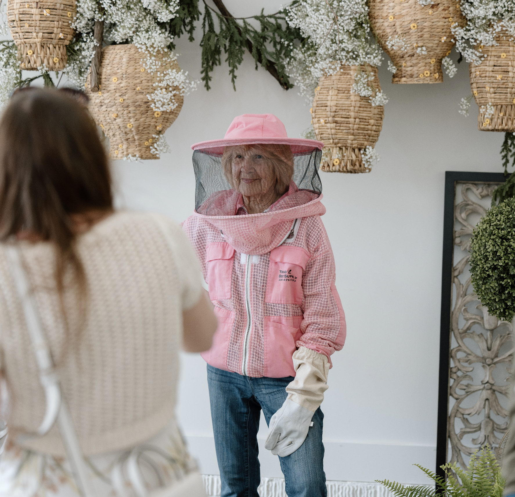 An elderly woman in a pink hat with a veil, pink shirt, and denim jeans, wearing beekeeping gloves and veil, standing indoors in front of hanging wicker bee skeps and white flowers, likely at a beekeeping or gardening event in Delaware, Ohio