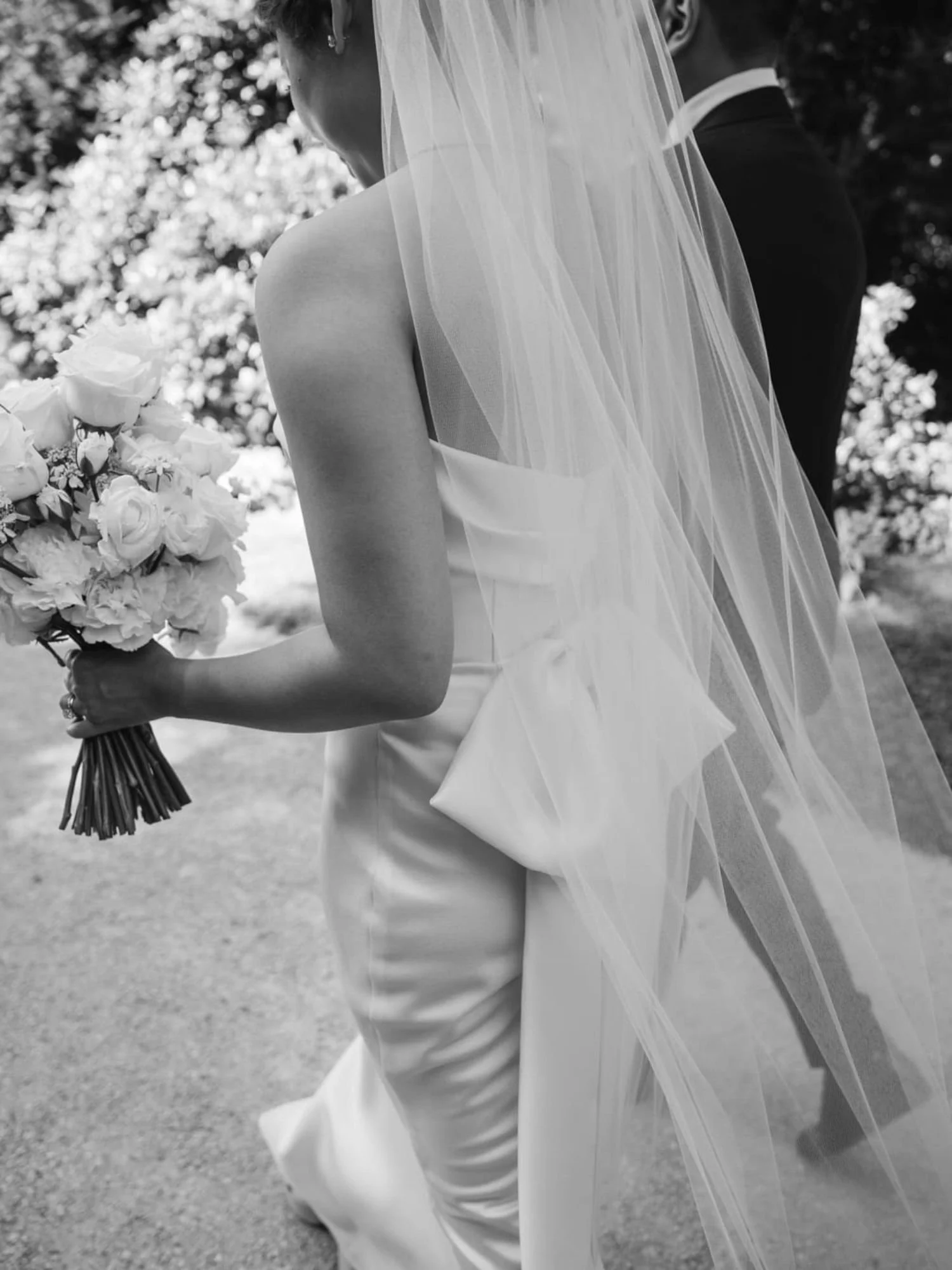 Braut läuft mit dem Bräutigam einen Weg entlang und hält Blumen in der Hand; sichtbar sind Kleid und Schleifendetail am Rücken.