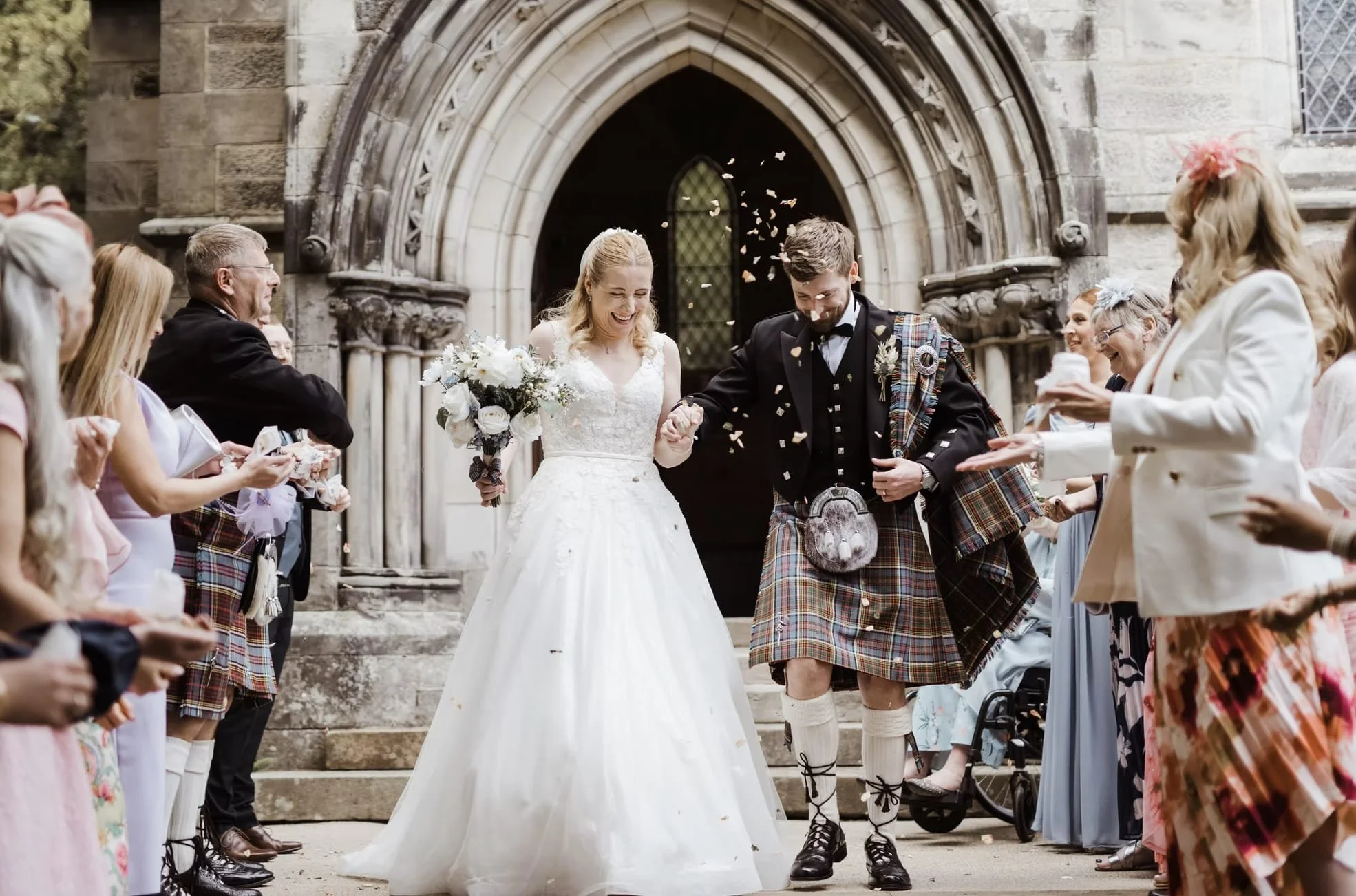 Brautpaar verlässt schottische Kirche, Gäste werfen Blüten, Bräutigam im Kilt