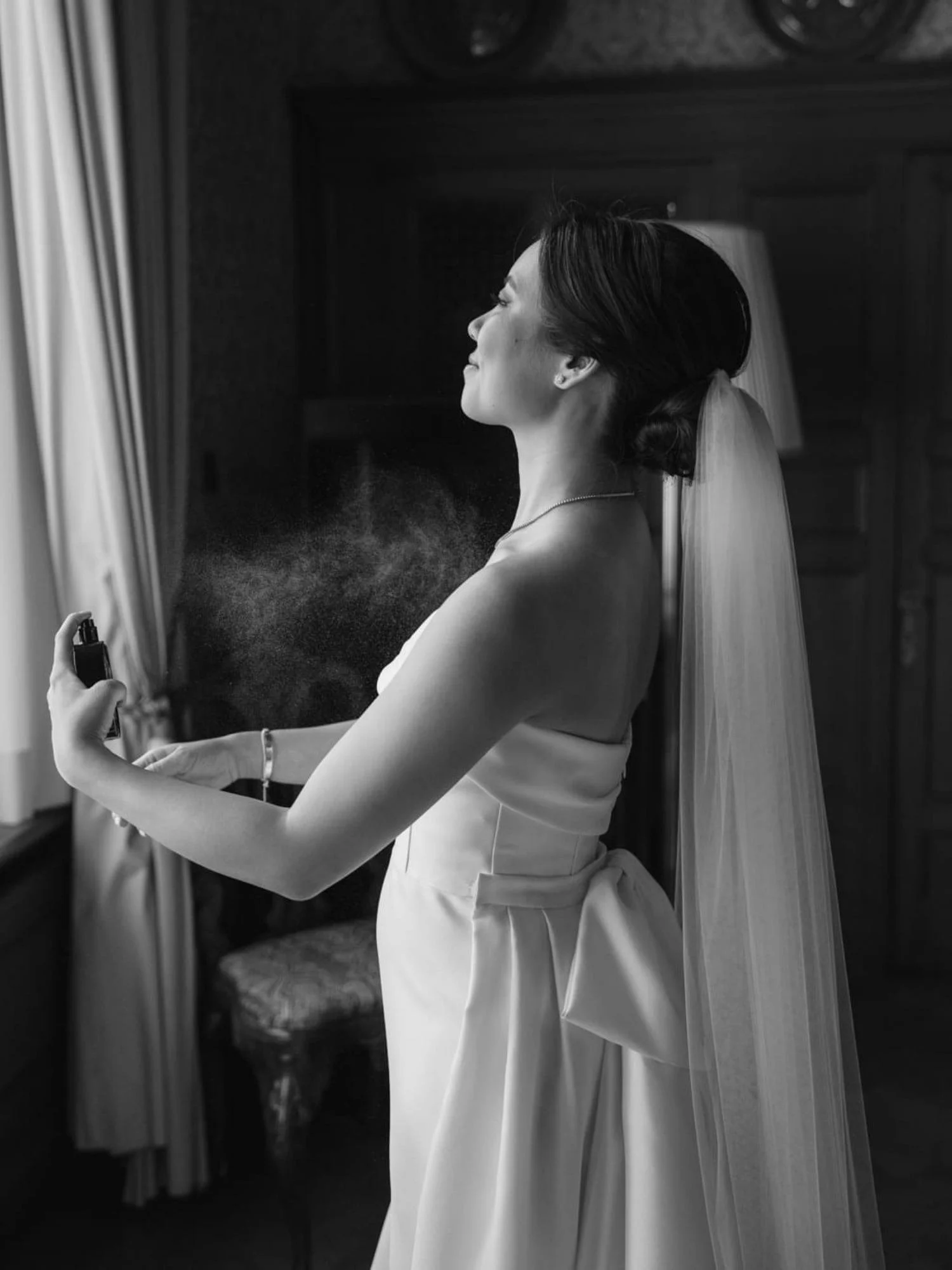 Braut sprüht sich kurz vor der Trauung ihr Parfum auf und steht angekleidet am Fenster der Hochzeitssuite im Schlosshotel Kronberg.