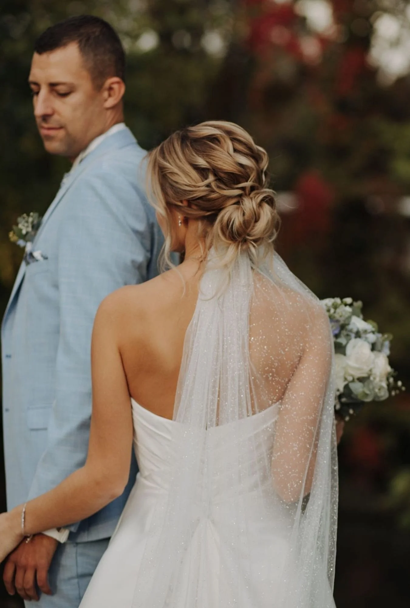 Braut mit Brautstrauß, Schleier und einem lockeren Haarstyling mit Bräutigam im Hintergrund