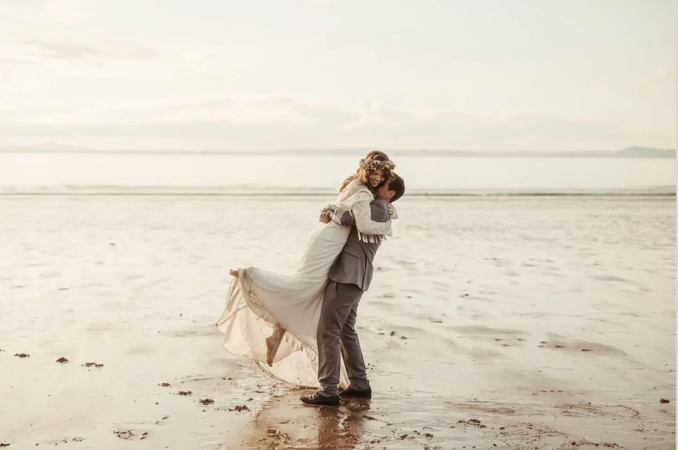 Hochzeitspaar am schottischen Strand, Braut mit Blumenkranz, Bräutigam hebt sie hoch, Sand und Wasser im Hintergrund