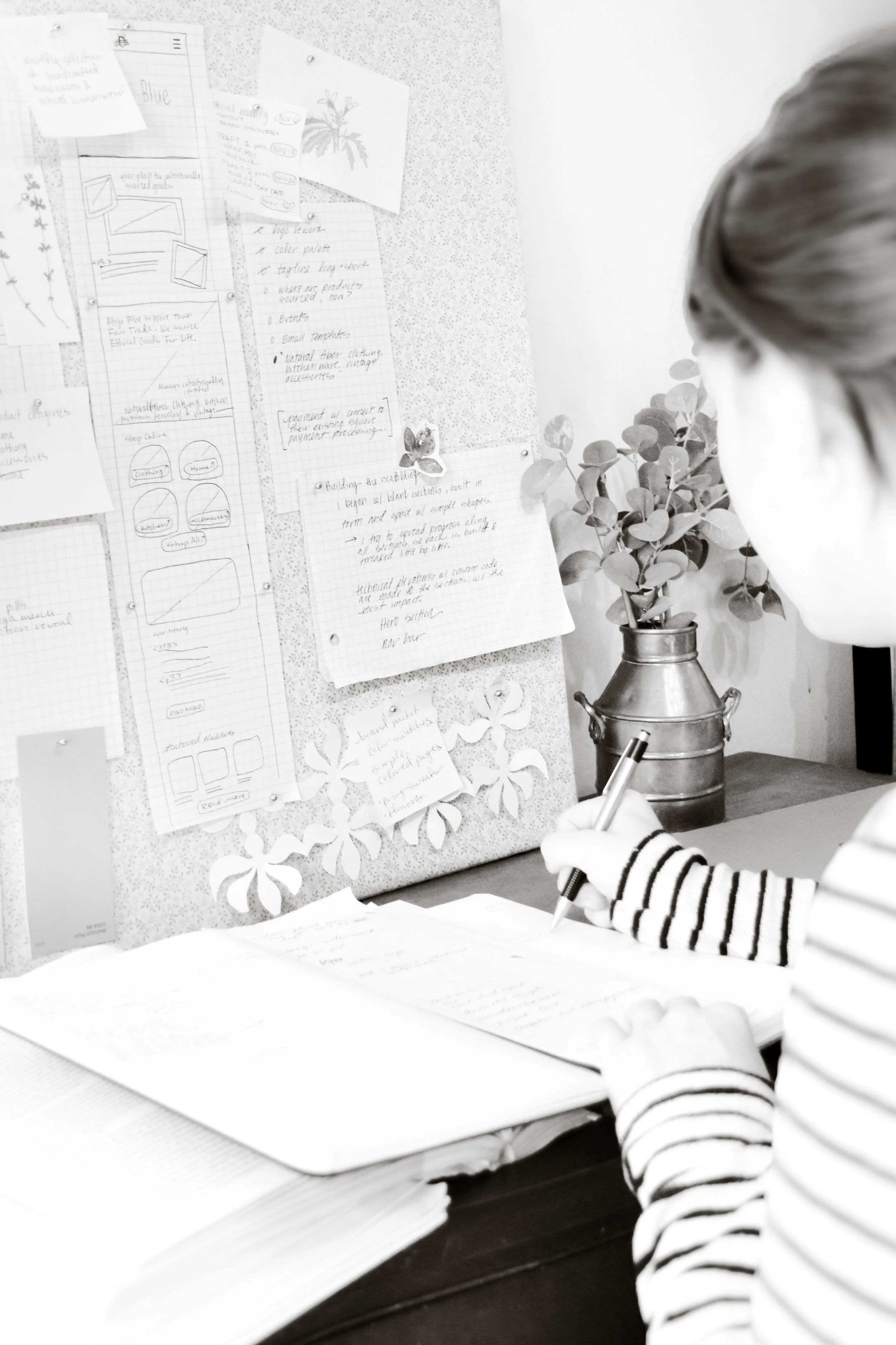 A person writing in a notebook at a desk, with papers and notes pinned on a bulletin board behind them, and a potted plant nearby.