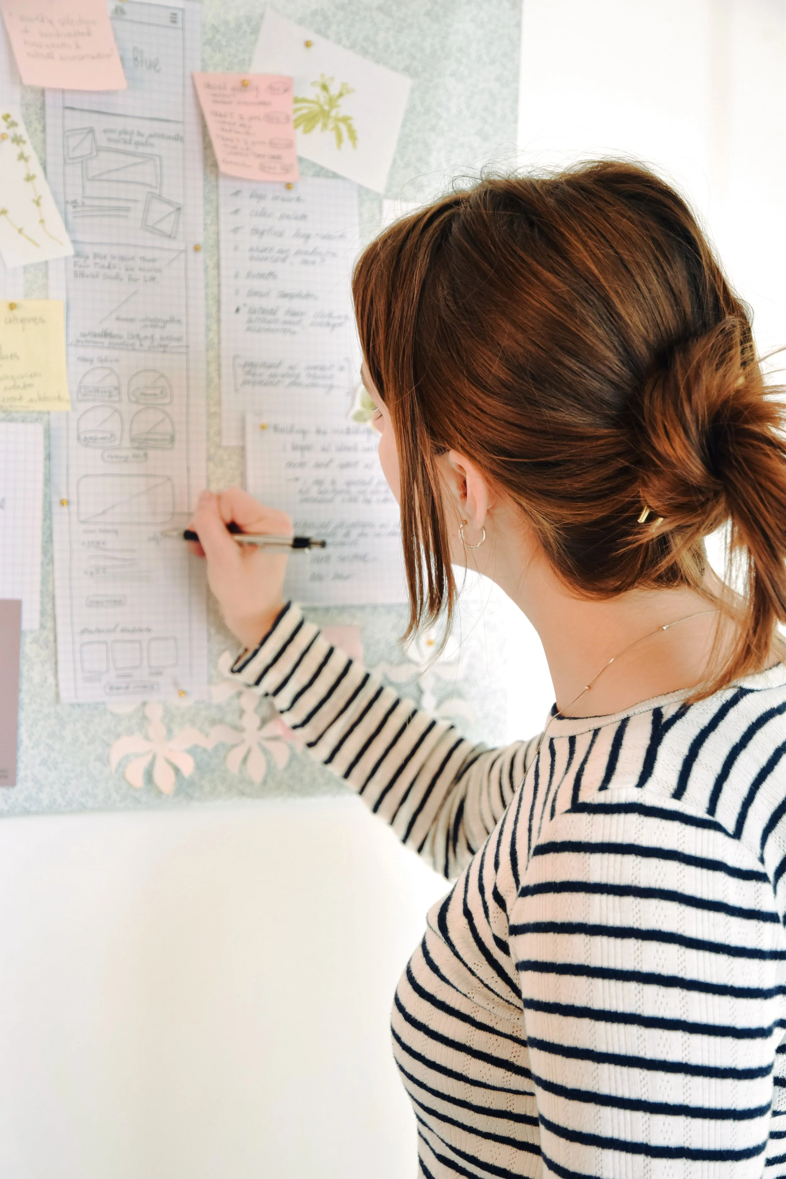 Woman with brown hair in a ponytail writing on a corkboard filled with notes and sketches.