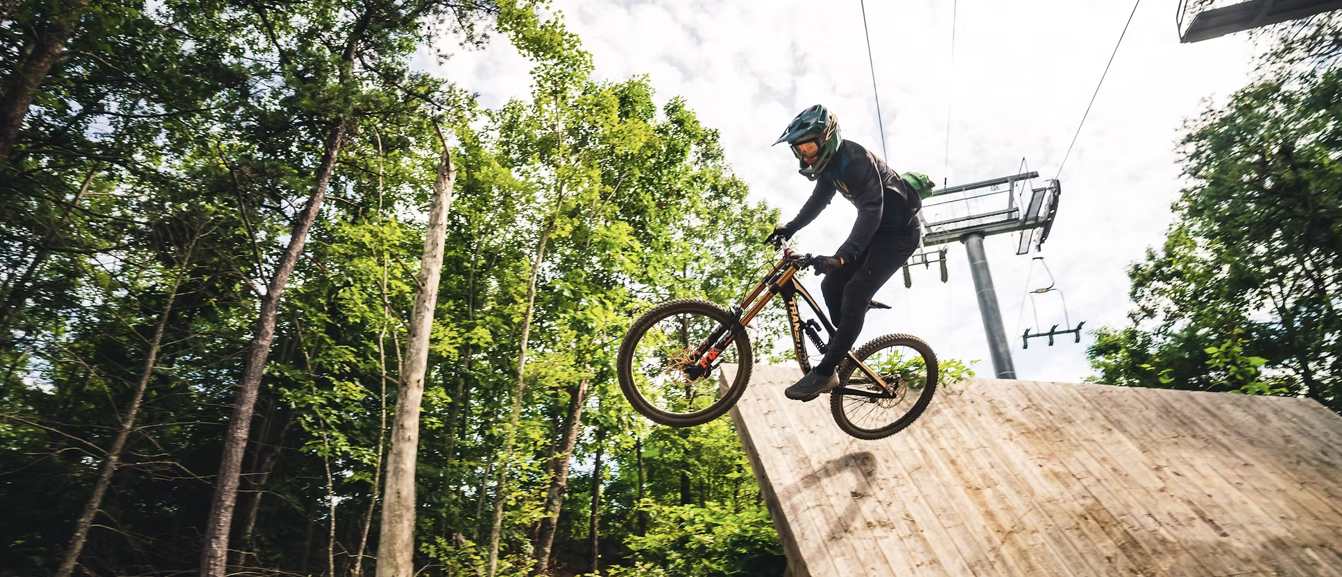 TAKEAIM Cycling owner riding a mountain bike wearing a helmet and black clothing rides off a ramp on a trail forested with green trees.