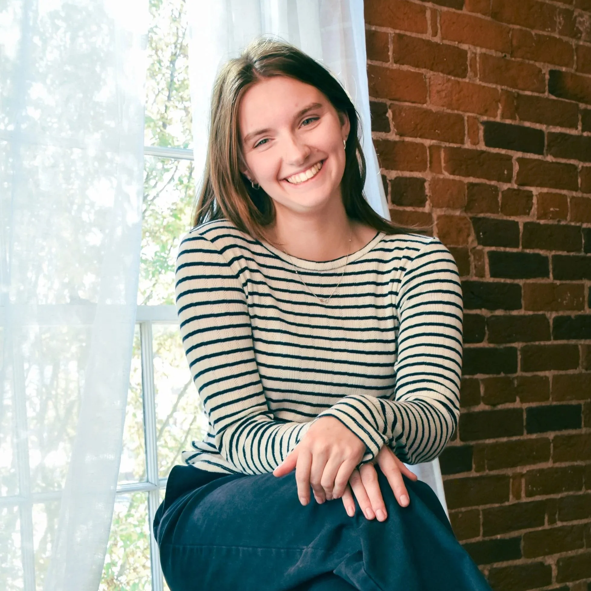A young woman with brown hair smiling, sitting by a window with light curtains and a brick wall behind her.