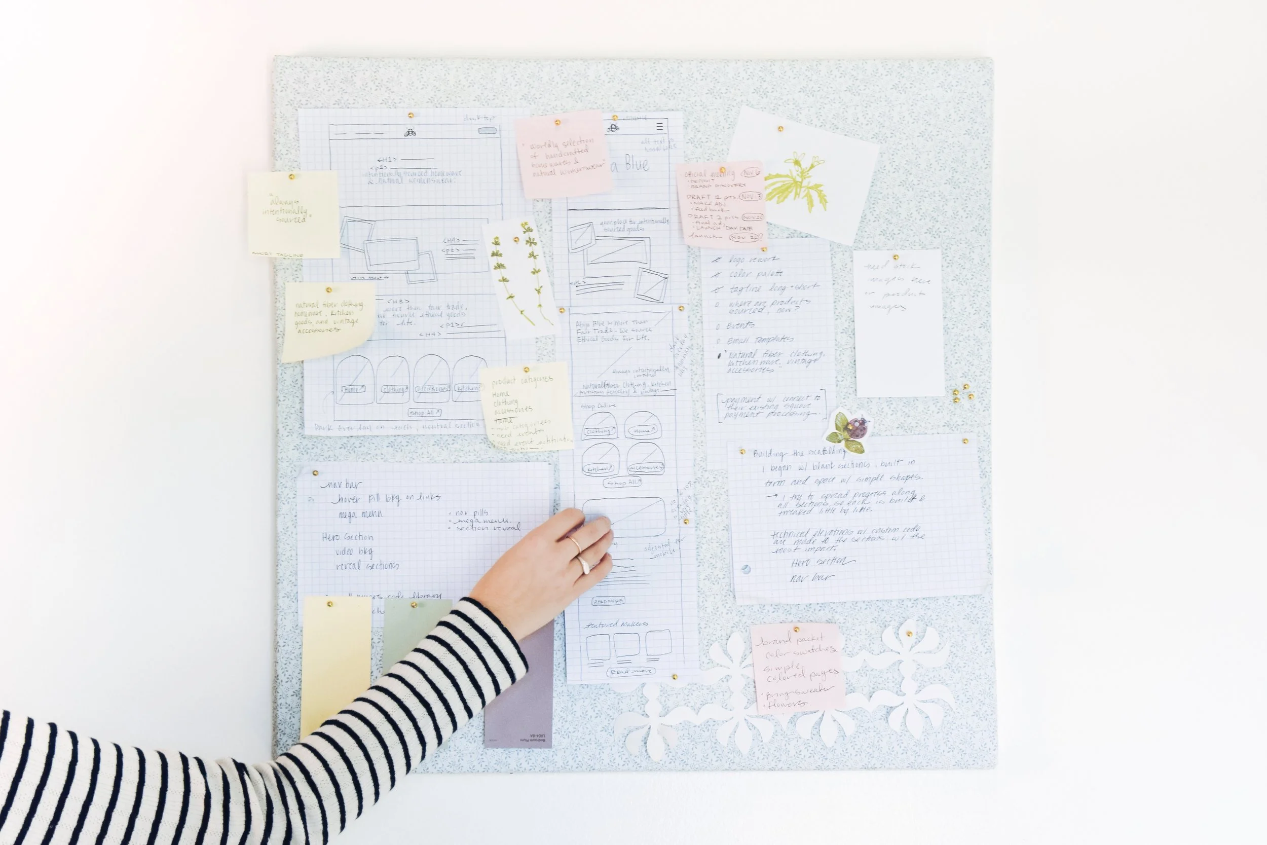 Person in a striped long sleeve shirt arranging notes and sketches on a decorative corkboard with sticky notes, drawings, and stationery.