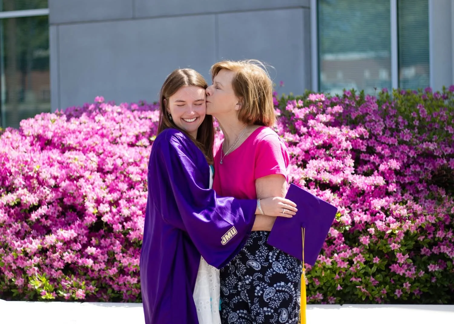 A graduation ceremony scene with a young woman in a purple graduation gown and an older woman, likely her mother, sharing a kiss on the cheek outdoors in front of pink azalea flowers, with a modern building in the background.