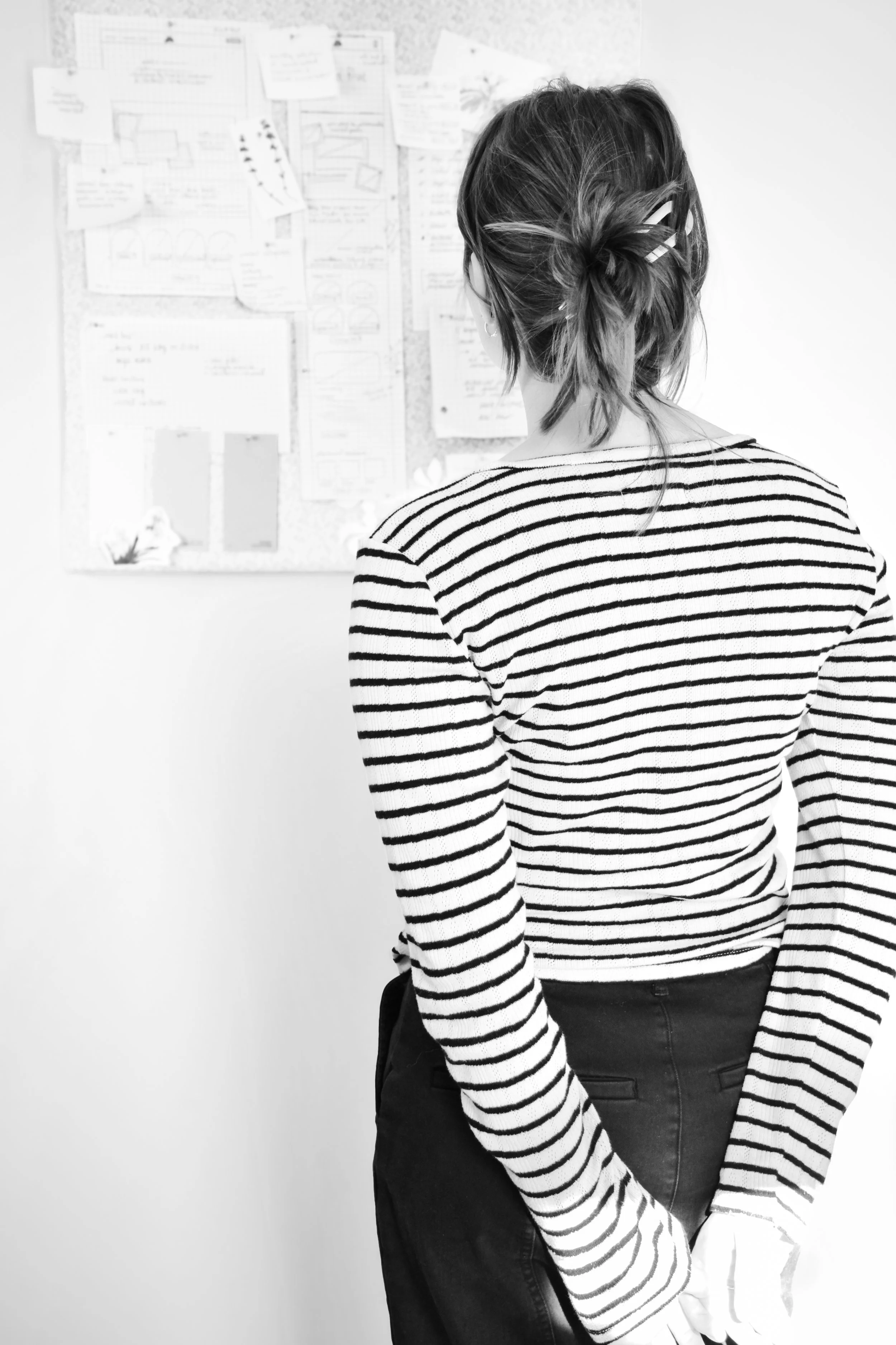 A woman with her back facing the camera, wearing a black and white striped long-sleeve shirt and black pants, looking at a bulletin board with papers and notes pinned on it.