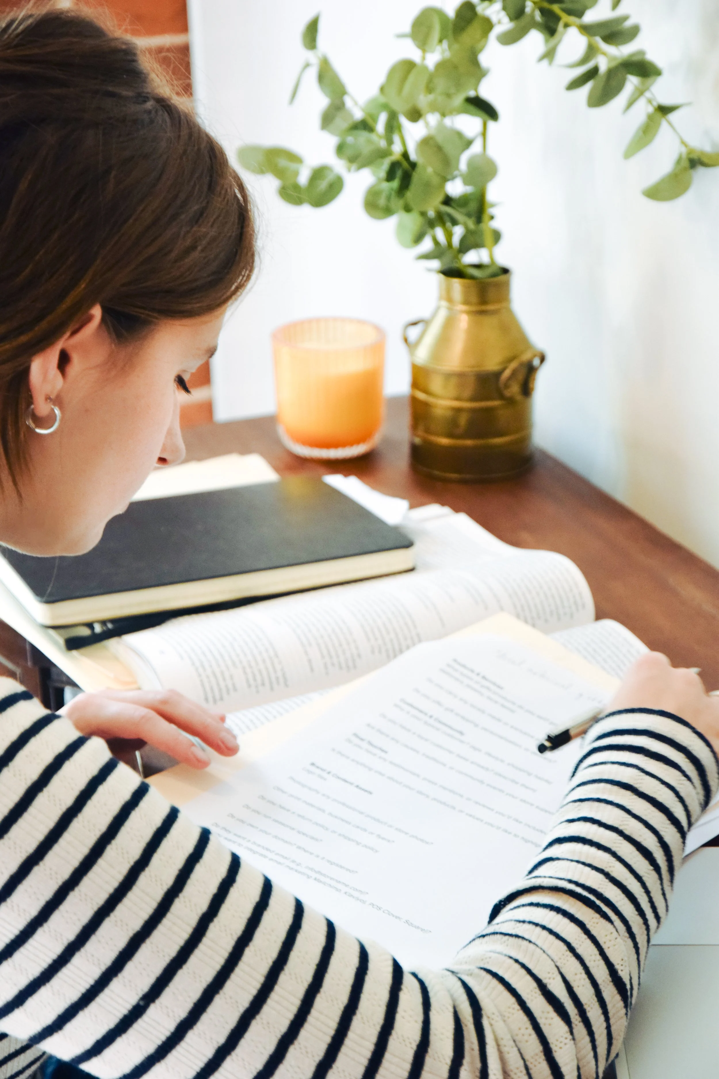 Person reading a book at a desk with notebooks, a plant in a gold vase, and a candle.