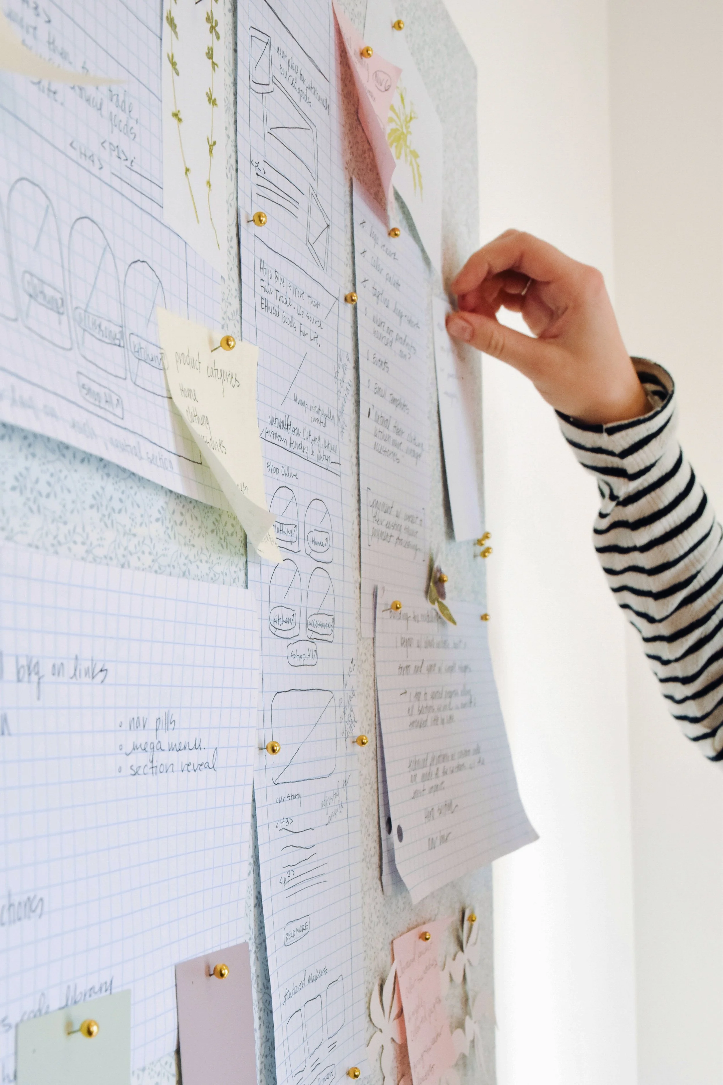 Person pinning a paper to a bulletin board filled with notes, sketches, and colorful paper pieces.