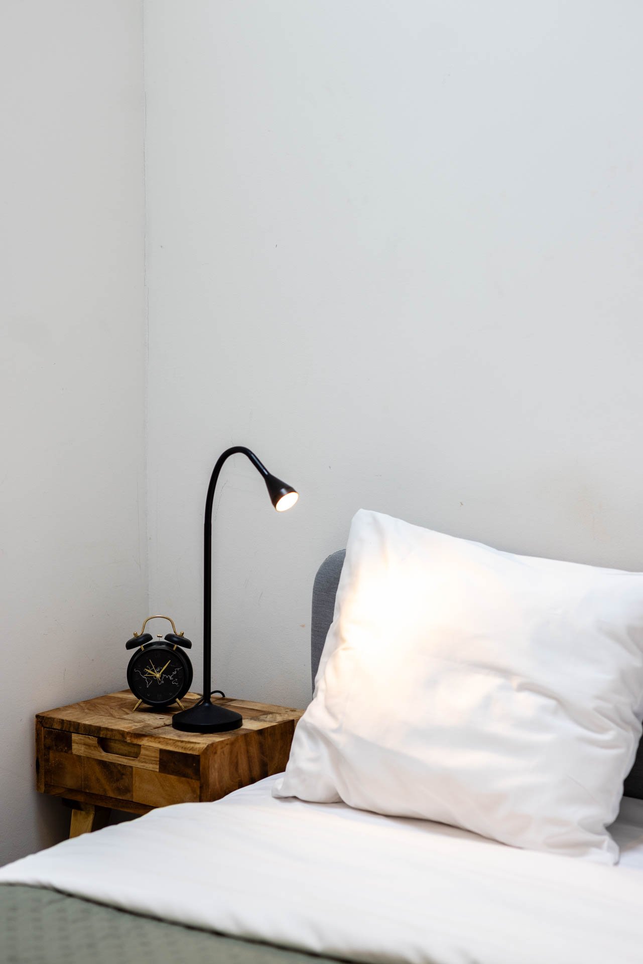 Slaapkamer met wit beddengoed, een houten nachtkastje, een zwarte bedlamp en een zwart wekker.