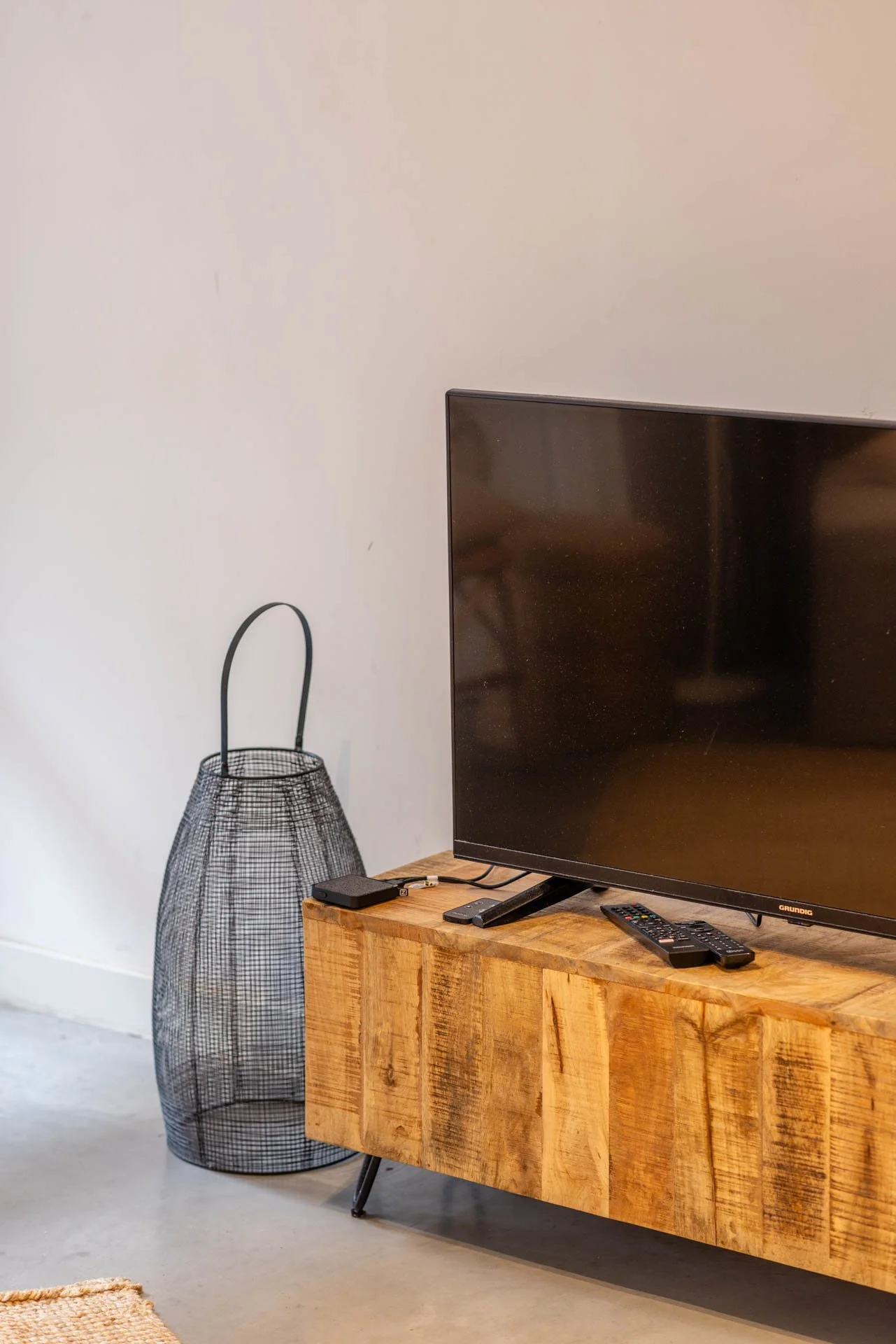 Een flatscreen-tv op een houten tv-meubel met afstandsbedieningen ernaast, en een metalen lamp op de vloer.