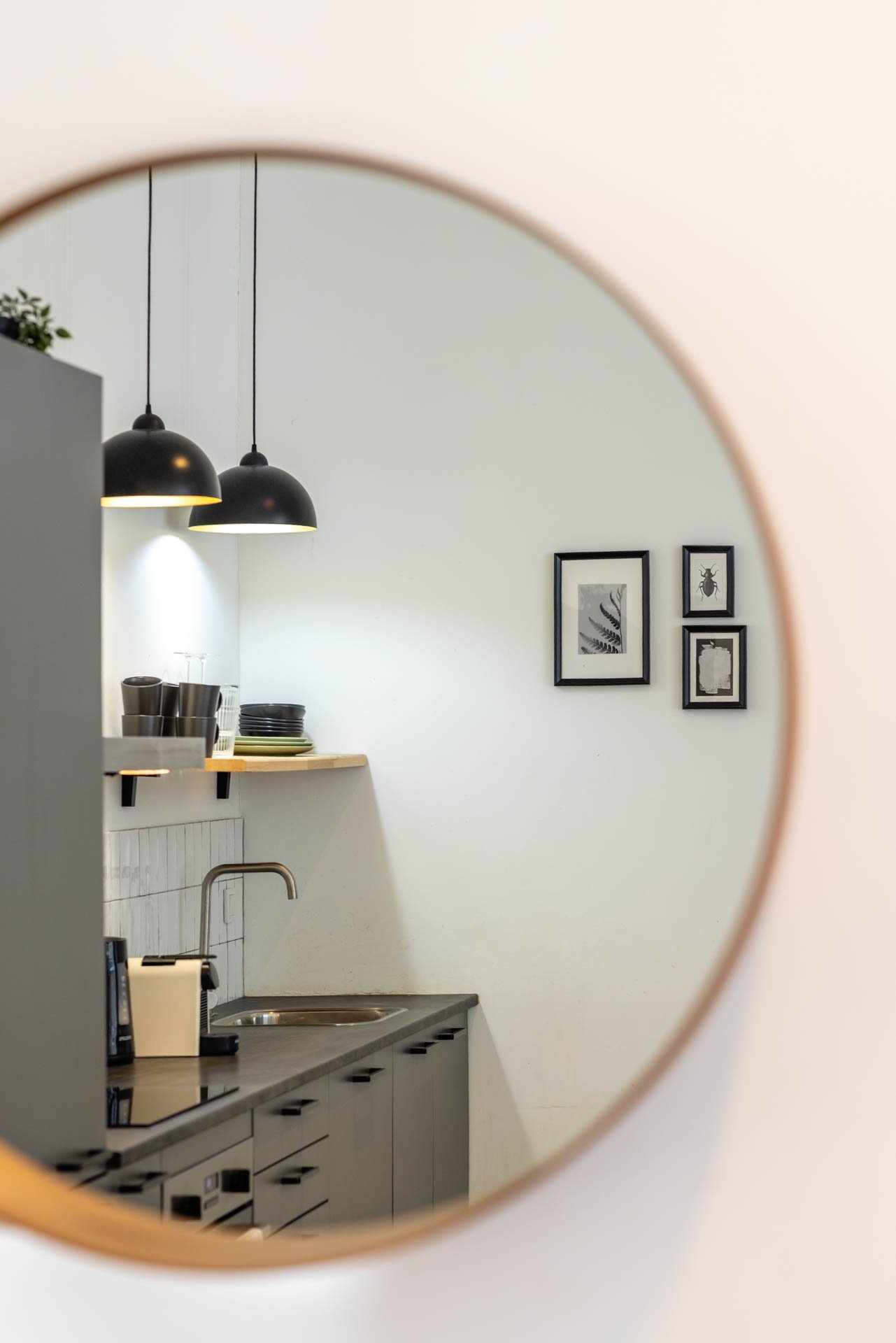 Kleine keuken met zwart en houtkleurig werkblad, vier zwarte plafondlampen, en vier zwart-witte kunstwerken aan de muur, zichtbaar via een ronde spiegel.