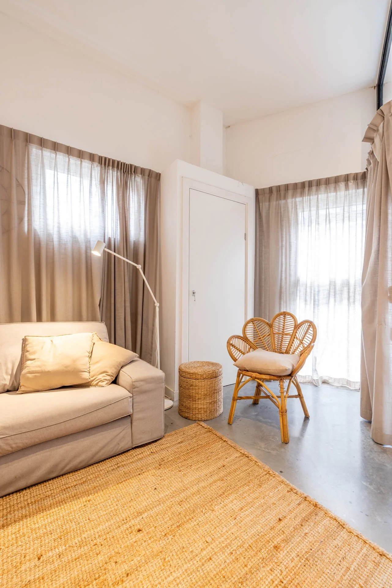 Een gezellige woonkamer met een beige bank, een leeslamp, gordijnen, een rieten stoel en een glazen deur met gordijnen.