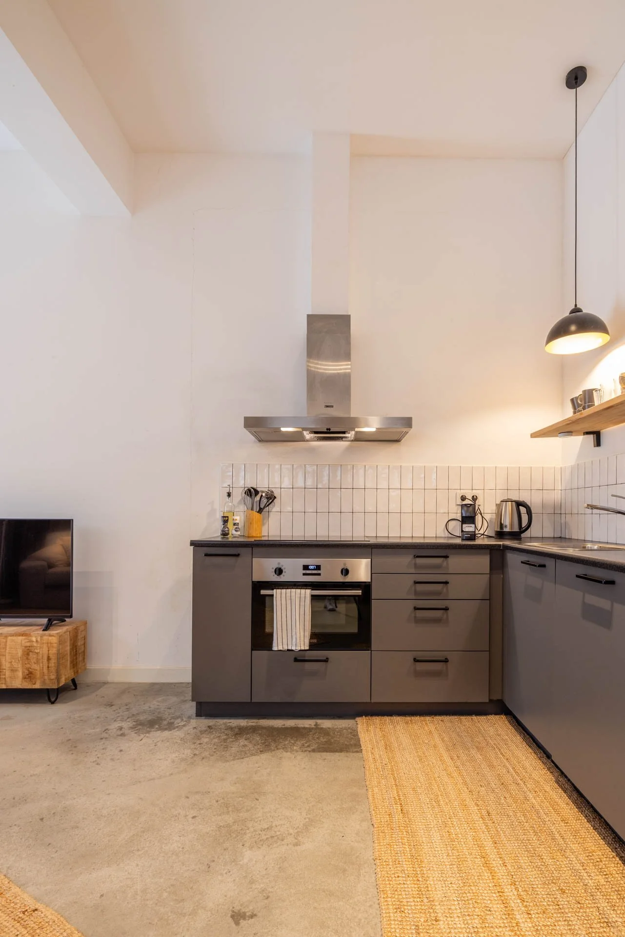 Moderne keuken met grijze kasten, een fornuis met oven, een afzuigkap, een houten muurplank met kopjes, en een ronde hanglamp. Aan de vloer ligt een beige mat en een tv staat op een houten rek.