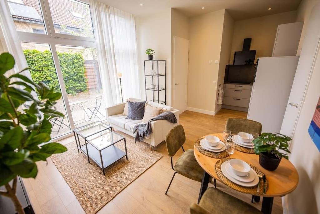 Een woonkamer met grote ramen, een witte bank, een glazen salontafel, een eettafel met borden en glazen, een tv, een open kast en planten.