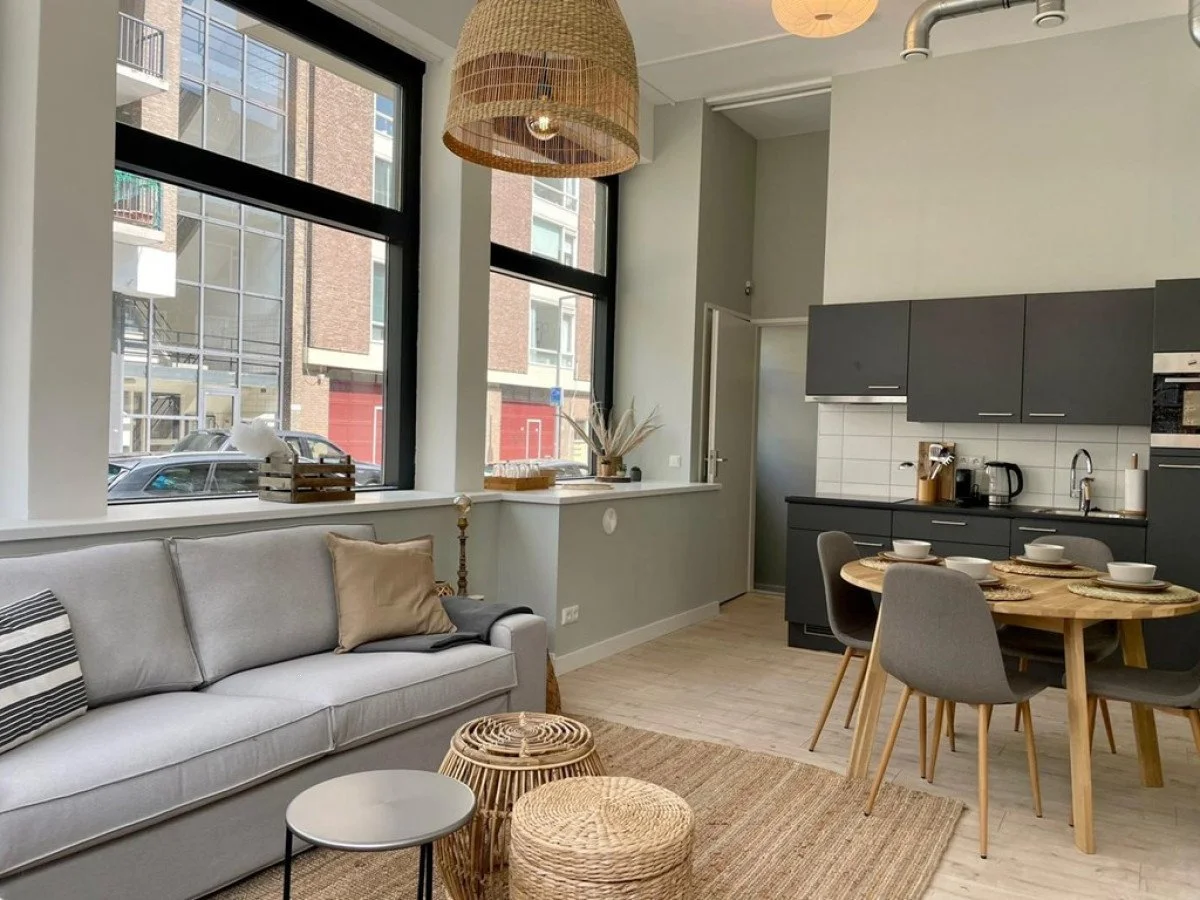 Een moderne woonkamer met grote ramen, een bank met kussens, een ronde eettafel met vier stoelen, een keuken met donkergrijze kasten, en decoratieve element zoals een hanglamp en vazen.