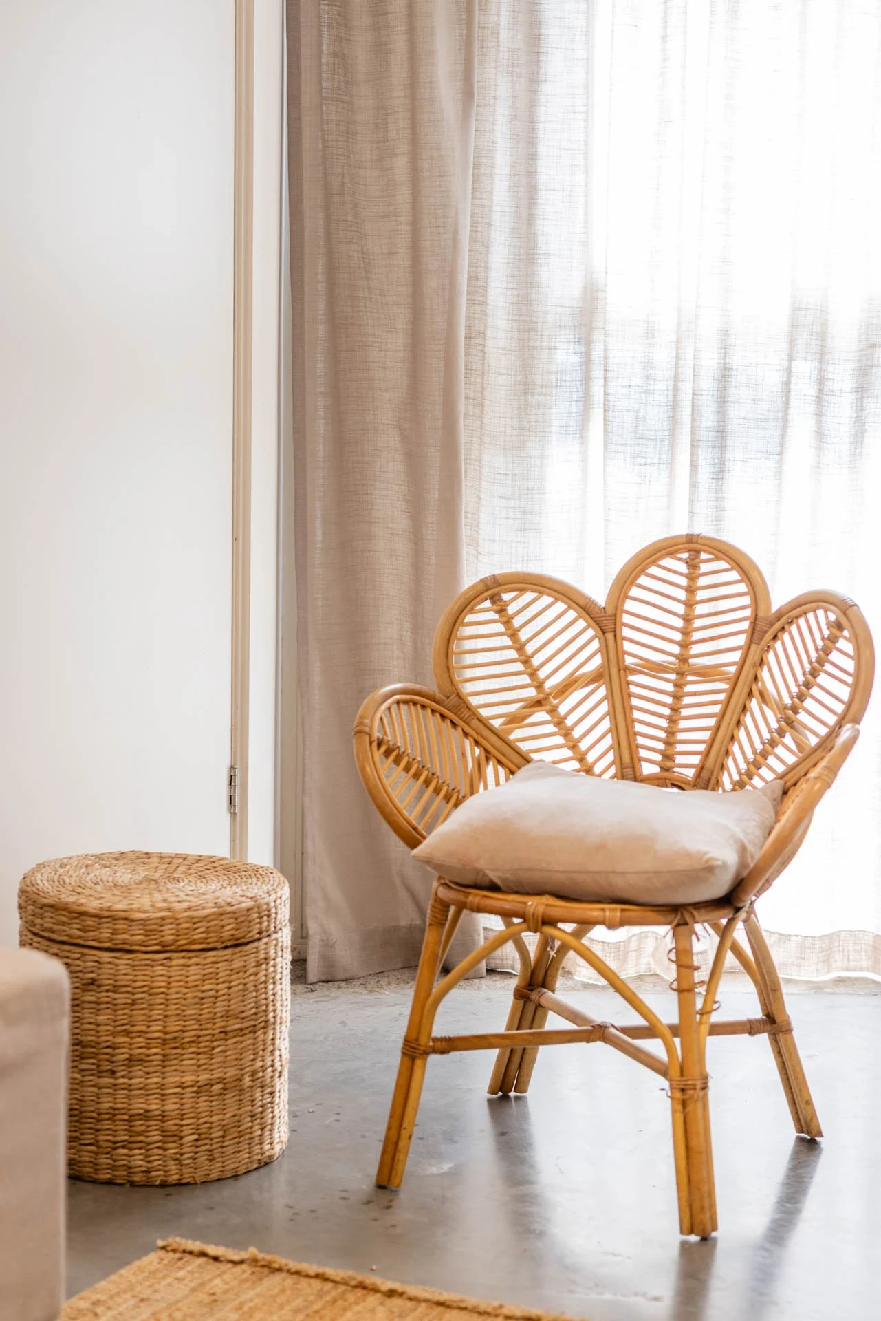 Een wicker fauteuil met een beige kussen, naast een ronde rieten krukje, in een lichte kamer met beige gordijnen.