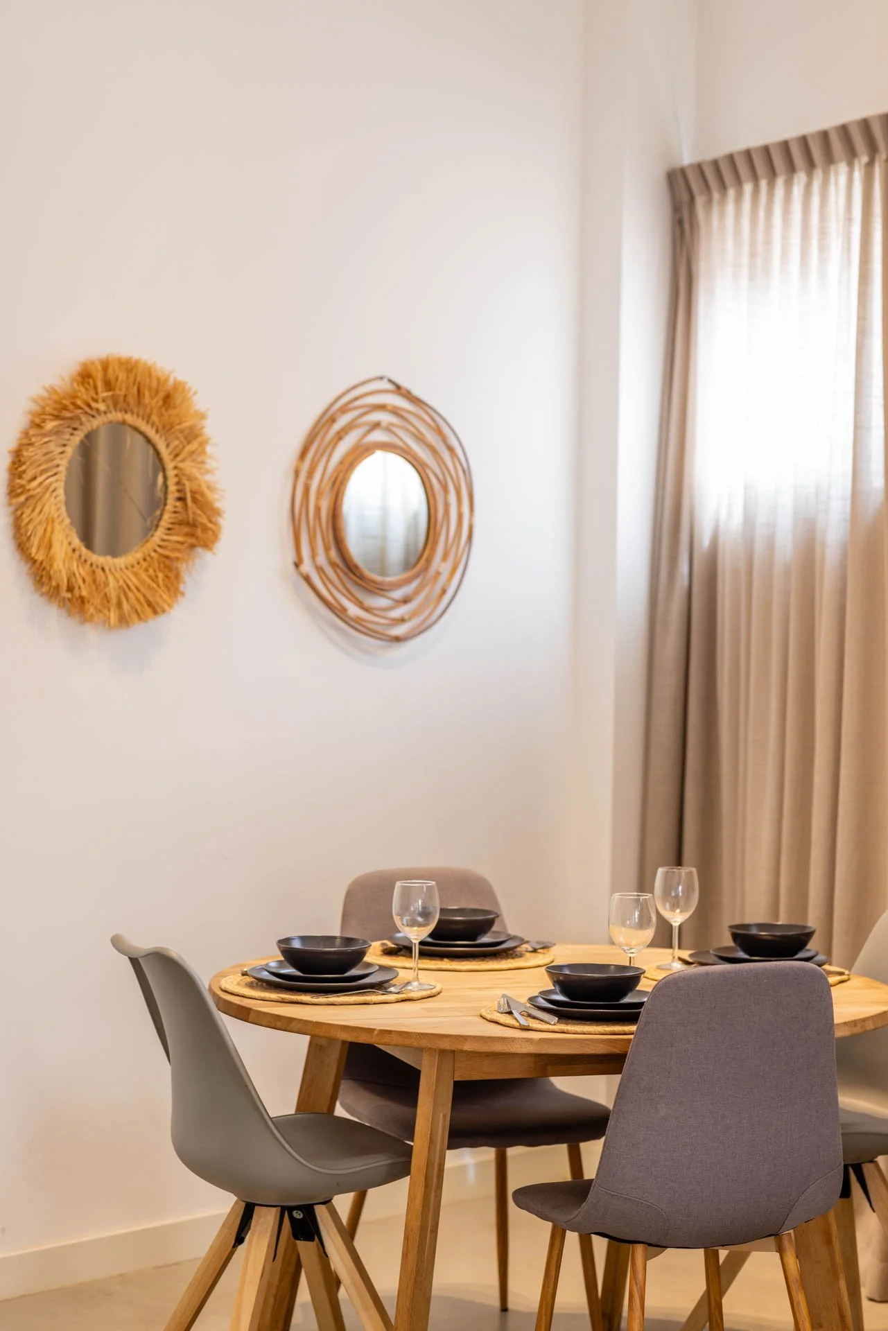 Een ronde eettafel met vier stoelen, klaar voor een maaltijd, met zwart servies en wijn glazen, in een kamer met beige gordijnen, witte muur en decoratieve spiegels aan de muur.