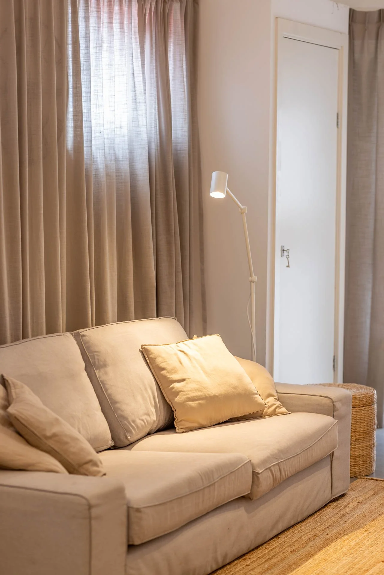 Een woonkamer met beige bank, sierkussens, gordijnen en een staande lamp.