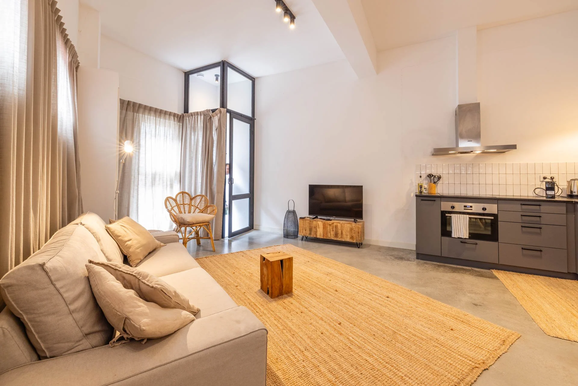 Modern open woonkamer en keuken met beige bank, houten tafel en tv, grote ramen met beige gordijnen, stenen vloer, keuken met grijze kasten, oven, afzuigkap en keukengerei.