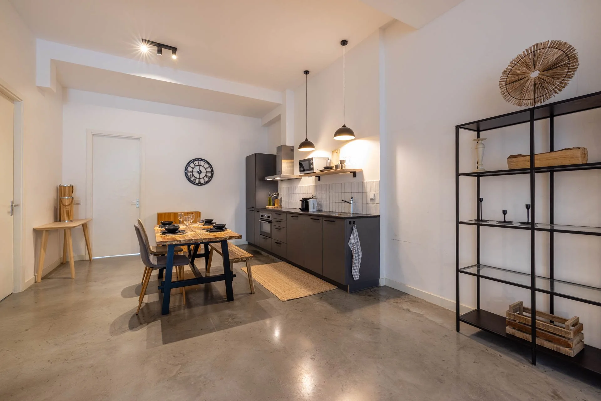 Eettafel met vier stoelen in een open keuken en woonruimte. De muur is wit en er hangt een grote kleine klok. De keuken heeft grijze kastjes en zwart werkblad. Aan de rechterkant bevindt zich een zwarte metalen plank zonder decoratie.