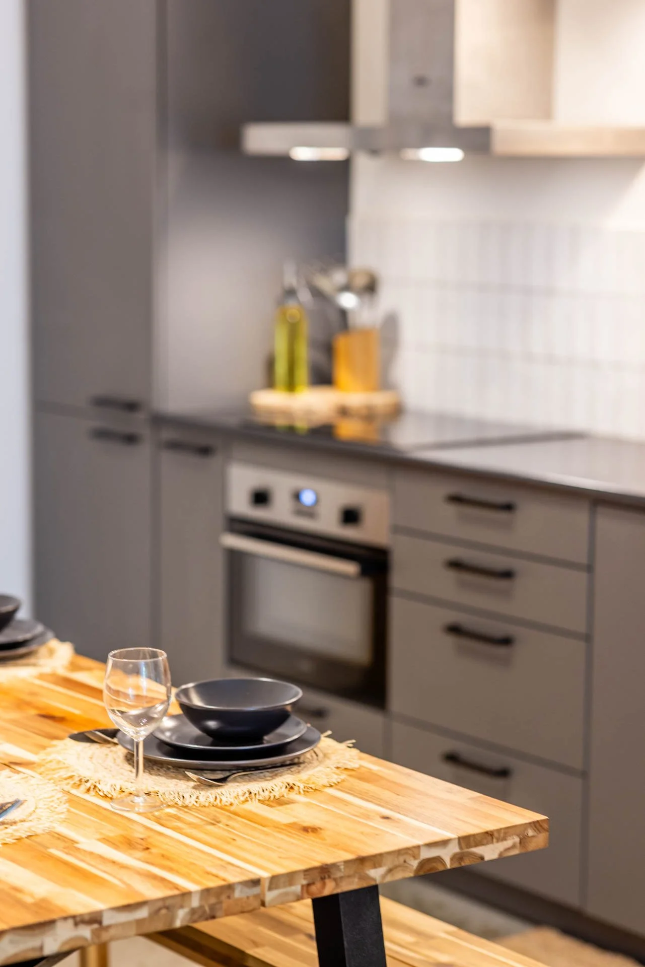 Een houten eettafel met zwart servies en een gevuld wijnglas in een moderne keuken.