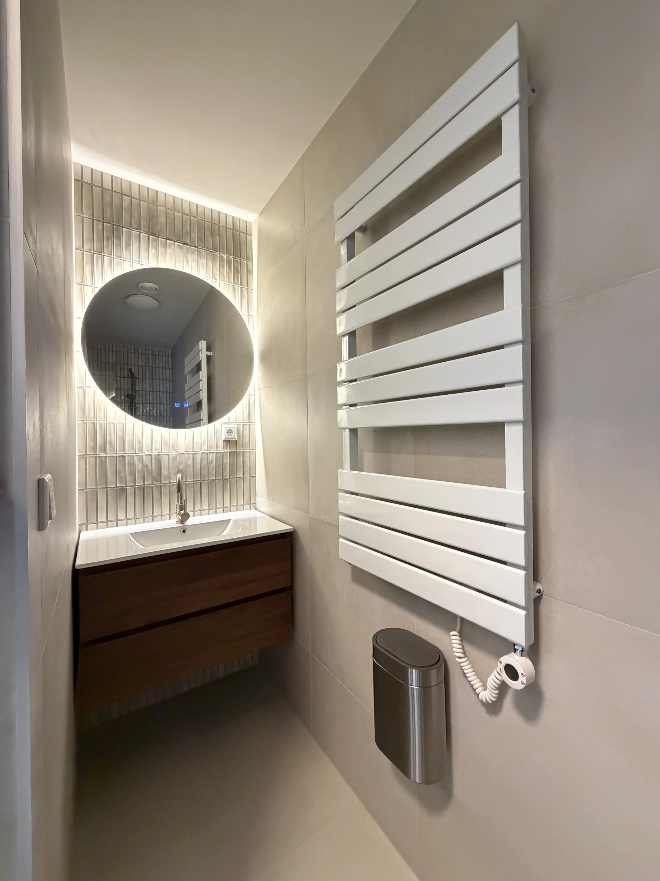 een moderne badkamer met een ronde muurspiegel, wastafel met lade onderhoek, en een witte radiator aan de muur.