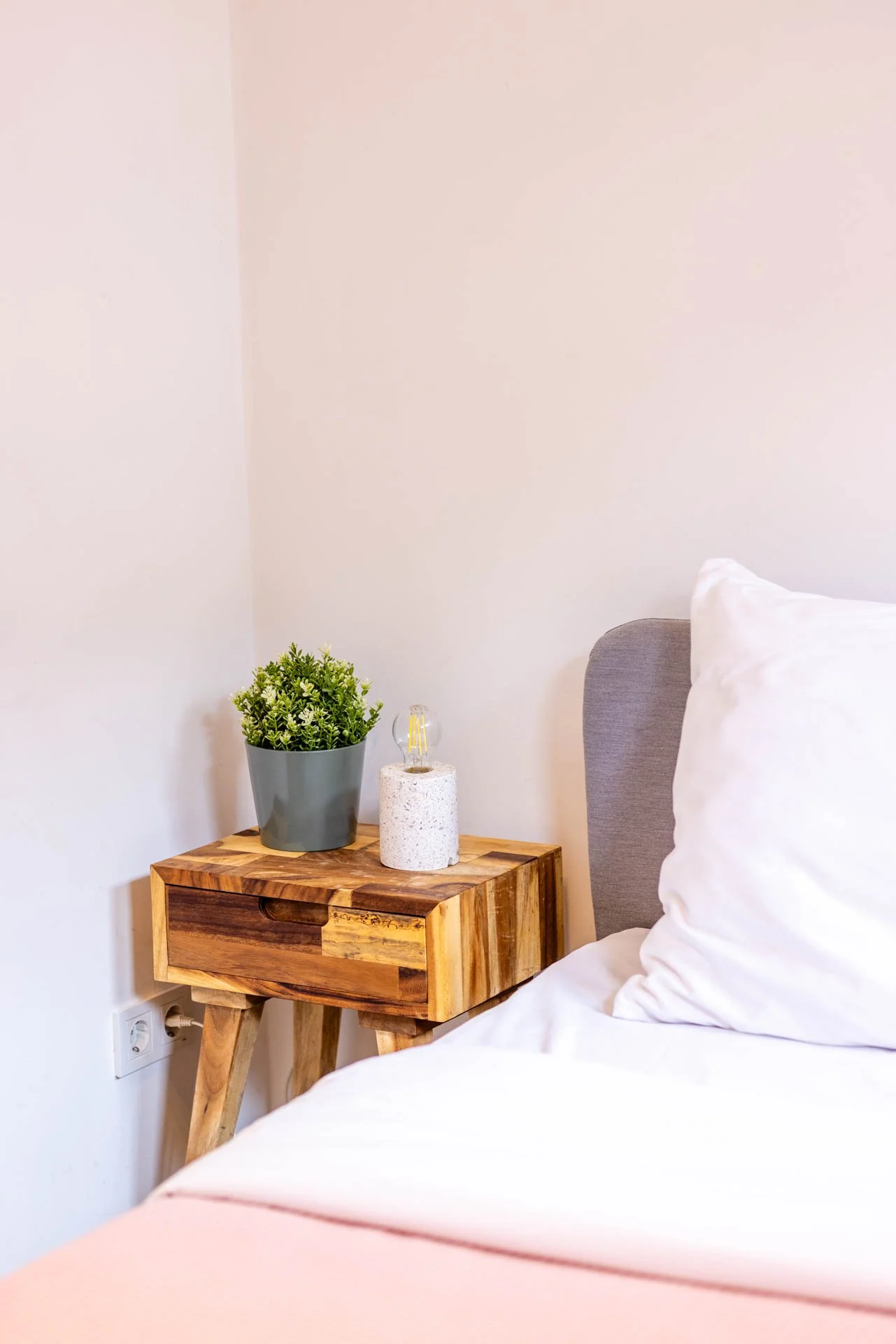 Een bed met witte lakens en een kussen, naast een houten nachtkastje met een groene plant in een grijze pot en een kleine lamp, tegen een witte muur.