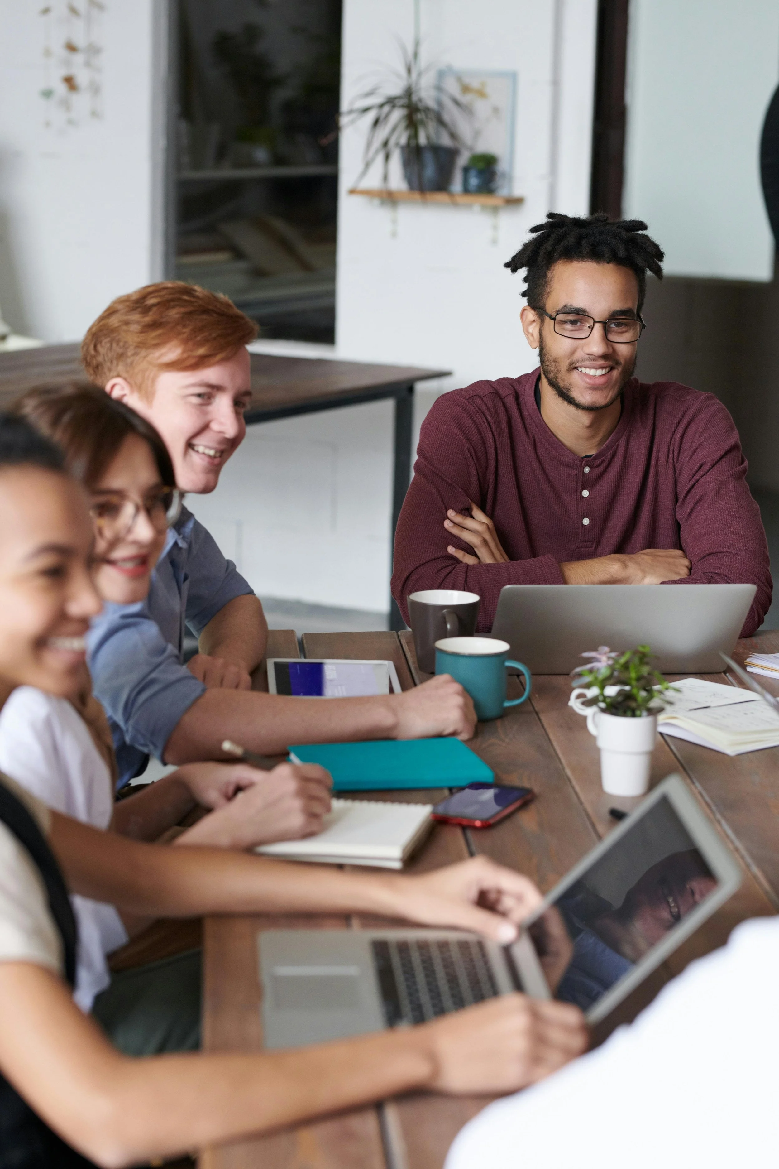 A group of young adults sitting at a conference table, smiling, with laptops, tablets, notebooks, and coffee mugs in a bright, modern office space.