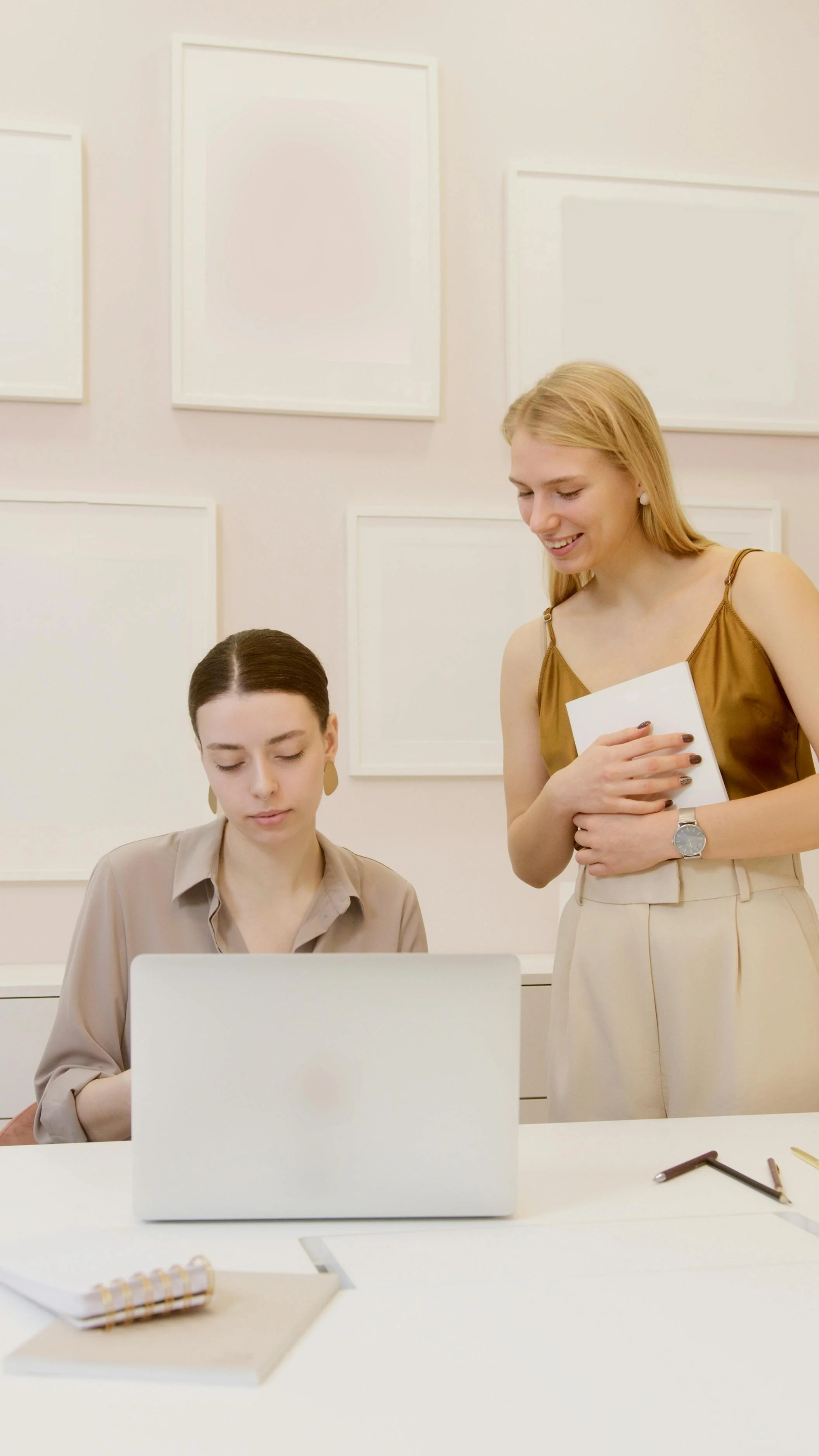Two women in an office, one sitting at a desk working on a laptop and the other standing, smiling, and holding some papers, with blank white frames on the wall behind them.