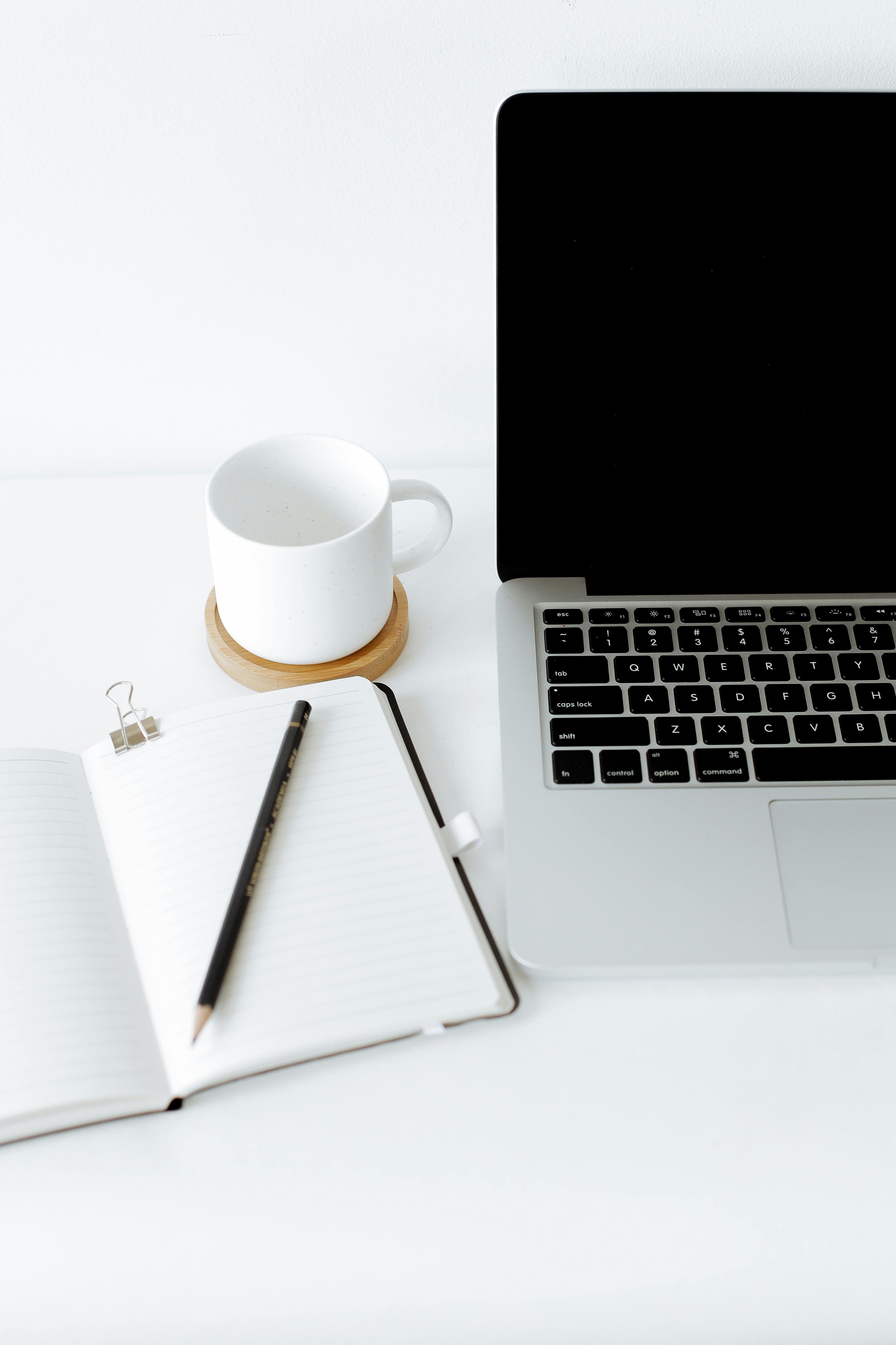 A workspace with a laptop, an open notebook with a pencil, a white mug on a wooden coaster, and a black paperclip on a white surface.