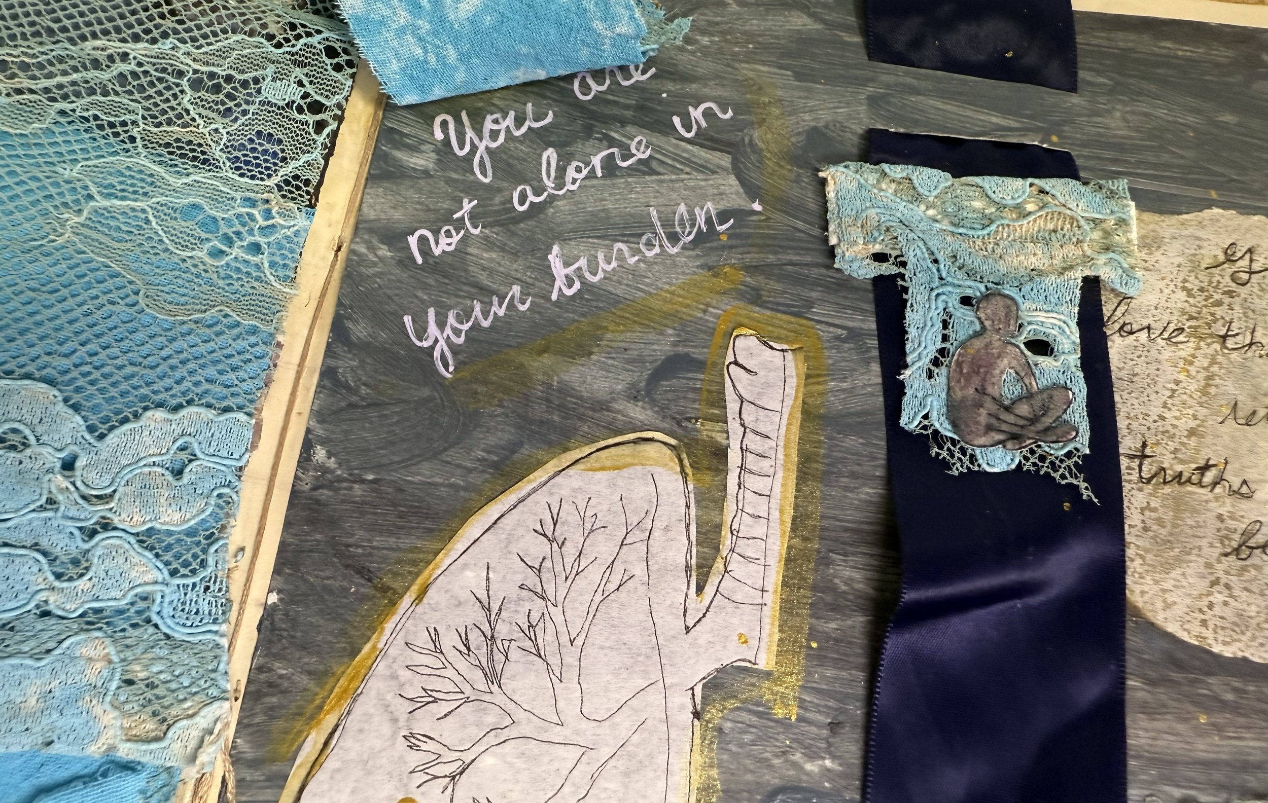 Various fabric and lace pieces, a handwritten note on a black surface with purple ink reading 'You are not alone in your struggle,' a simple outline drawing of a person with trees, and a ribbon featuring fabric with lace and a small cutout of a human figure.