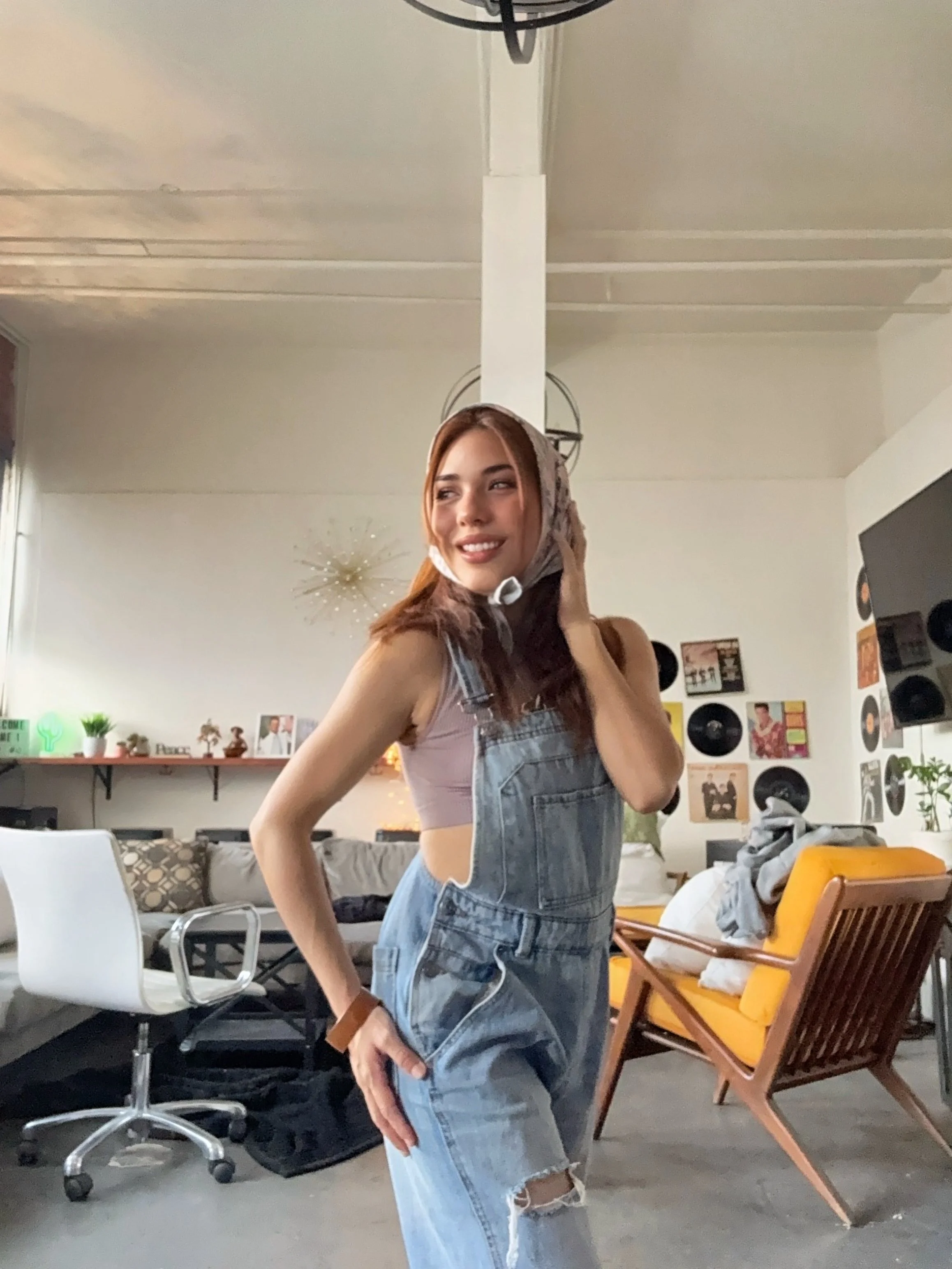 A young woman with a scarf on her head smiling and posing in a living room with eclectic decor, including a white office chair, a yellow armchair, and framed album covers on the wall.