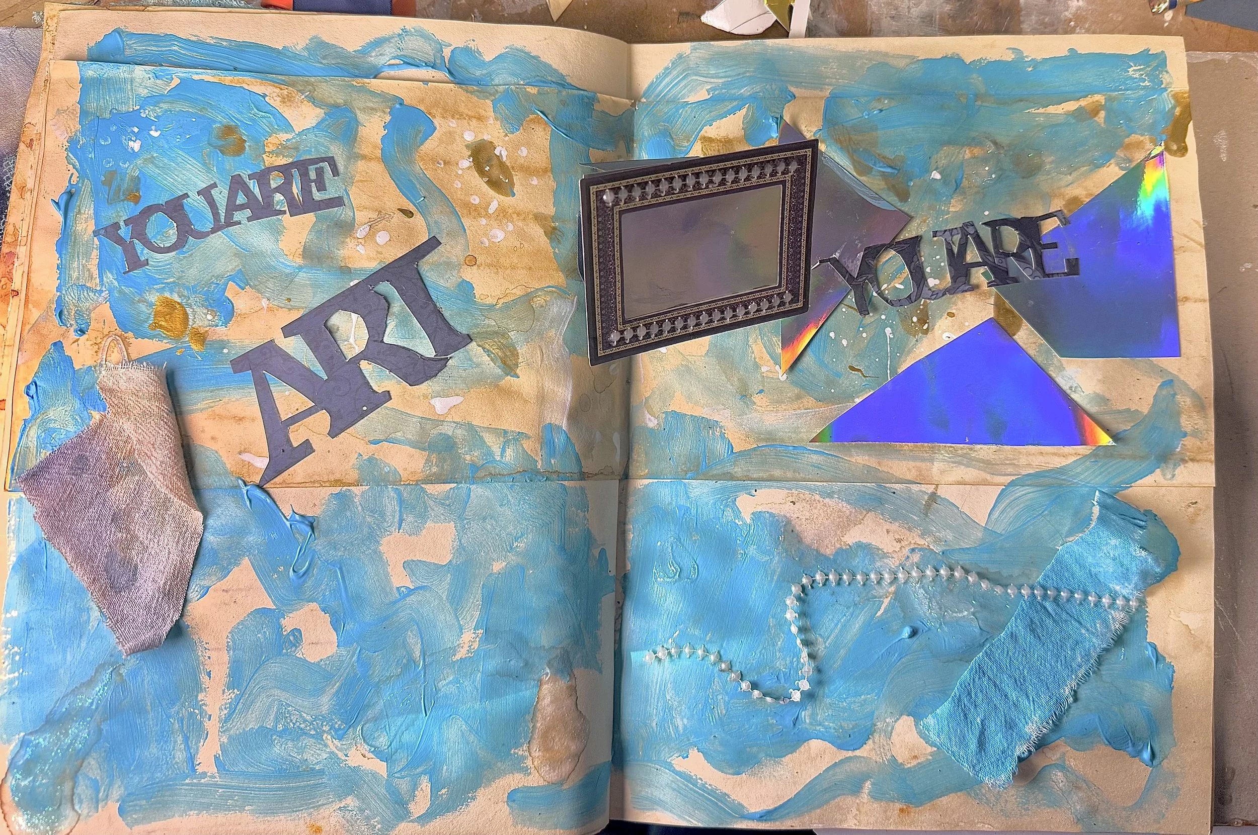 An art journal page with blue painted swirls, cut-out words saying 'You are' and 'Unique', holographic triangles, a picture frame, a piece of fabric, a pearl necklace, and gold splatters.