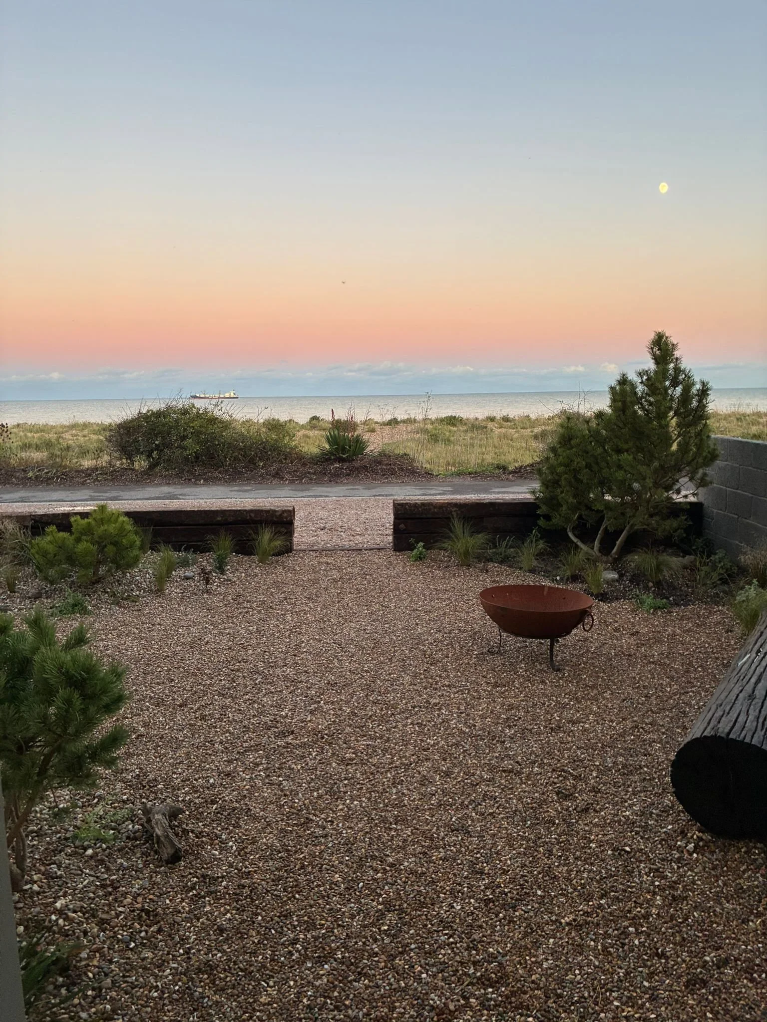 A gravel patio with a fire pit, small trees and bushes, overlooking a beach and ocean during sunset with a colorful sky and a visible moon.
