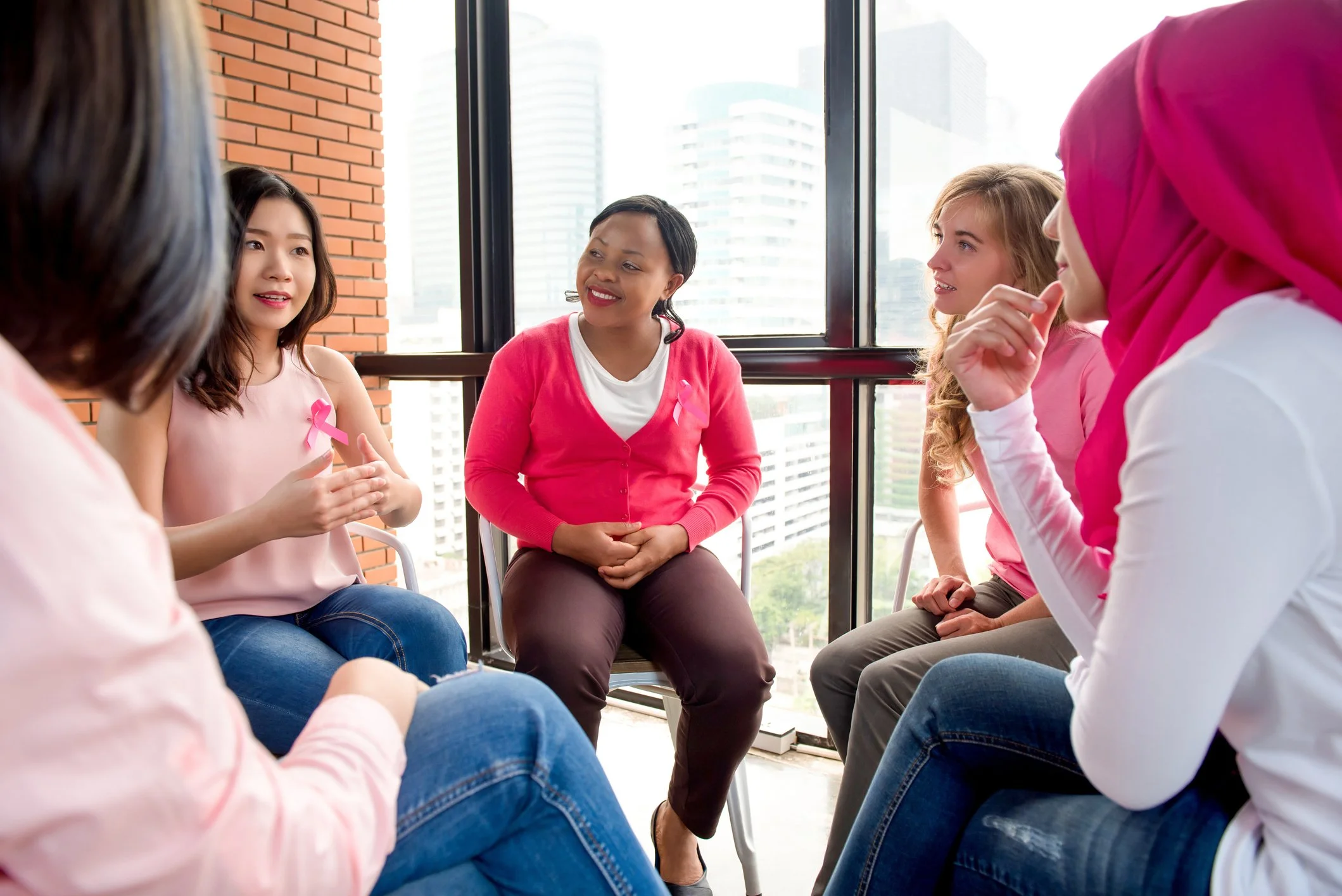 A group of women sitting in a circle in a bright room with large windows, participating in a support or discussion group. Some are wearing pink in support of breast cancer awareness.