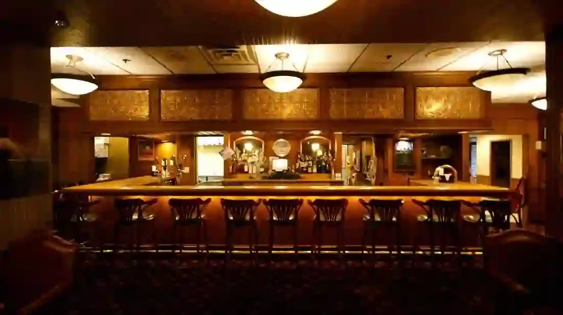 Dimly lit bar with wooden counters, bar stools, and back bar with bottles, glasses, and a cash register, in a cozy setting.