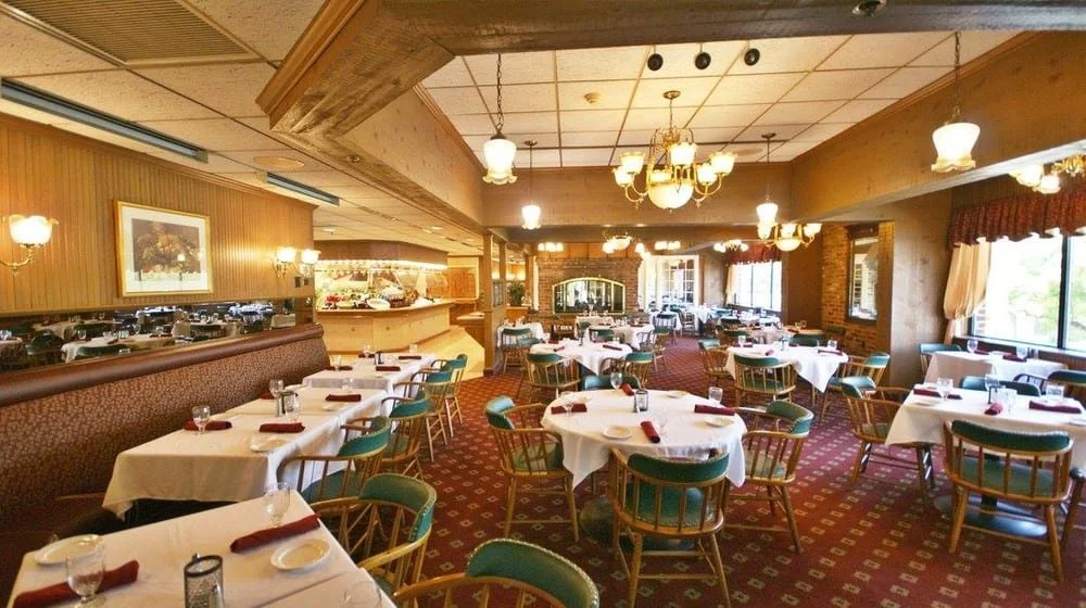 Dimly lit bar with wooden counters, bar stools, and back bar with bottles, glasses, and a cash register, in a cozy setting.