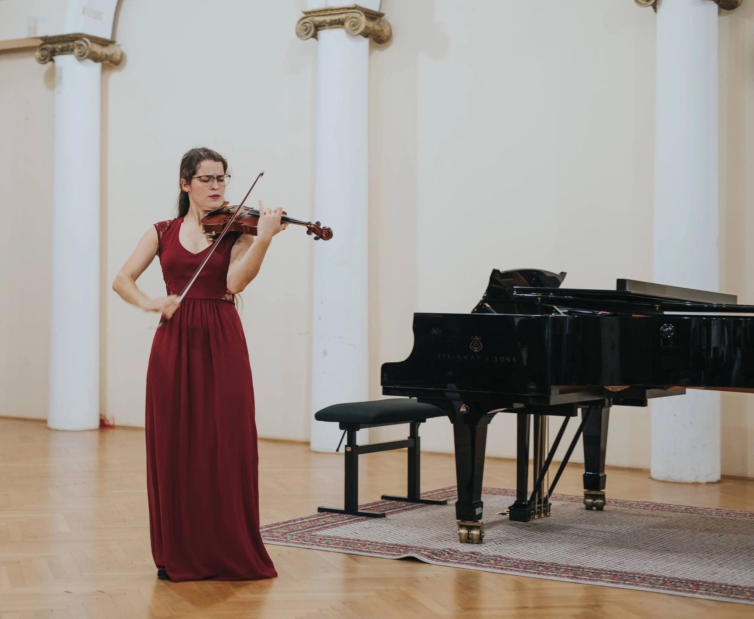 A woman wearing a long red dress playing a violin next to a grand piano in a room with white walls and columns.