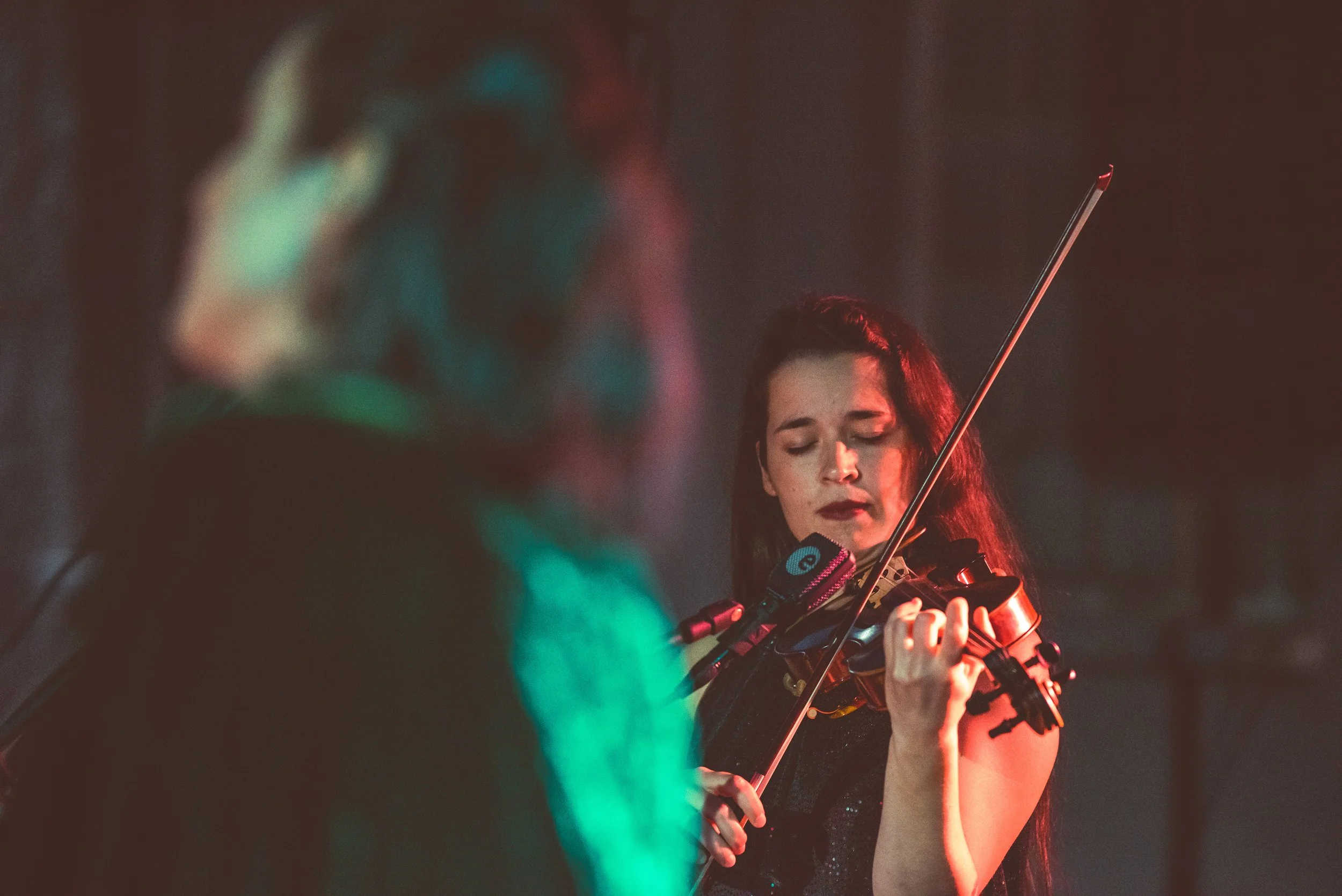 A woman playing the violin with a bow, with her eyes closed and a focused expression, in a dimly lit setting with colored lighting.